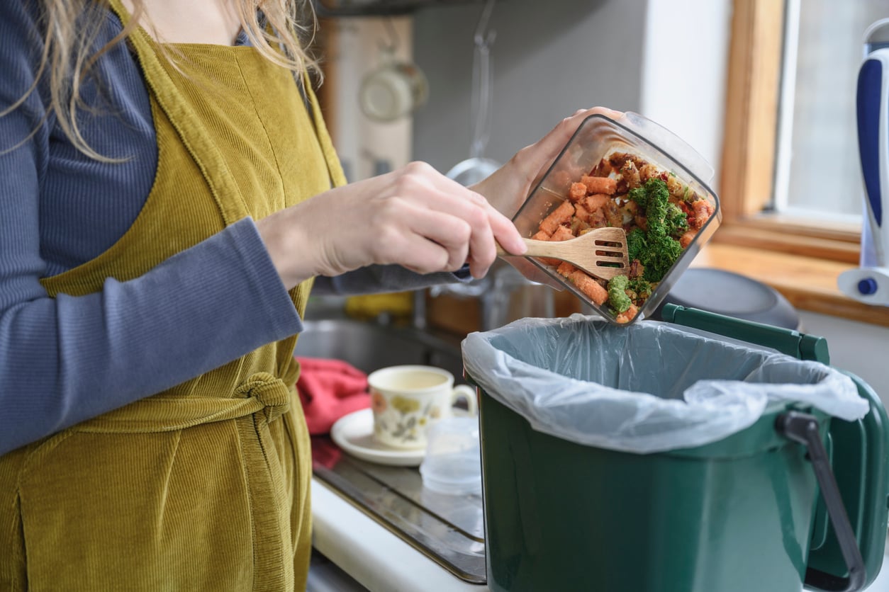 A person wearing a green apron, mindful of their grocery shopping habits, scrapes food scraps from a glass container into a green compost bin in a kitchen.