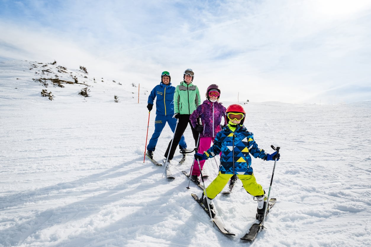Four people wearing colorful ski outfits and helmets stand together on a snowy ski slope, smiling at the camera under a bright, partly cloudy sky—enjoying the kind of winter adventure that makes memberships worth paying for.
