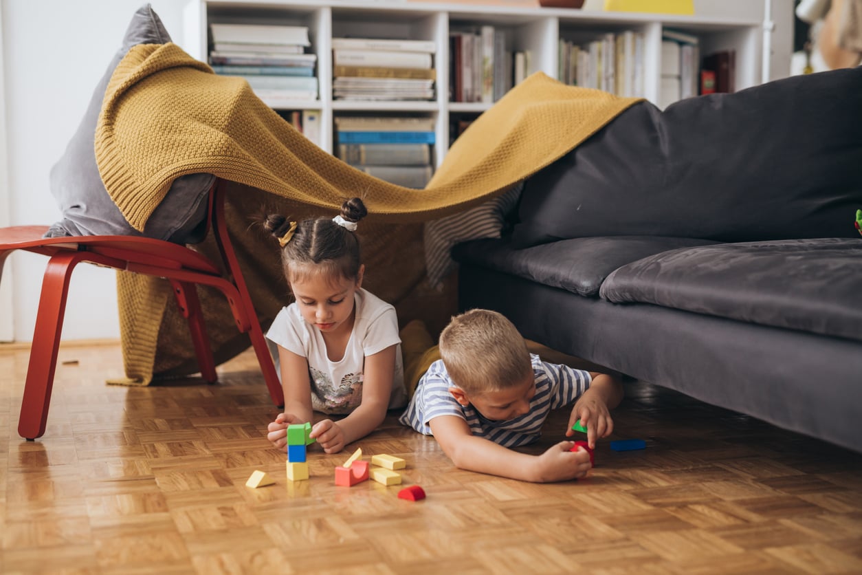 Two young children play with colorful blocks on a wooden floor in front of a makeshift fort made from a sofa and a yellow blanket, with shelves and books in the background.