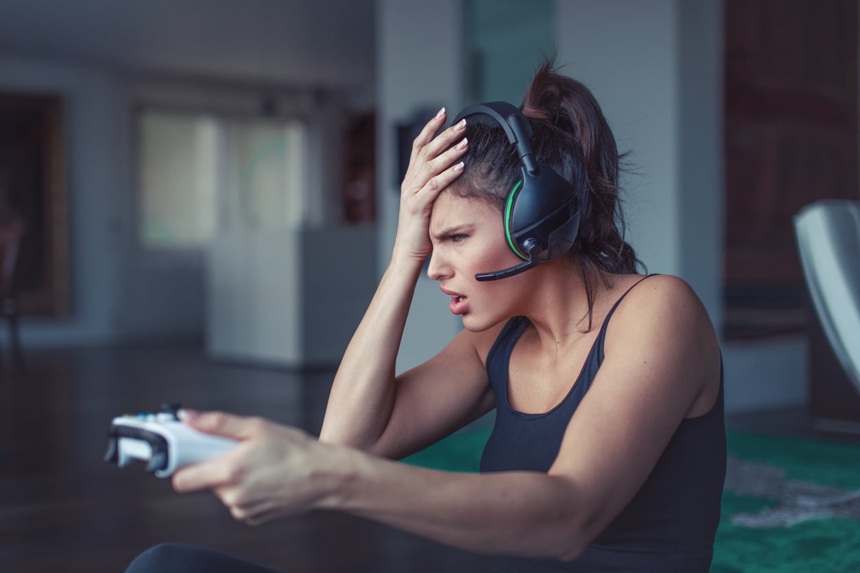 A woman wearing a headset holds a video game controller and looks frustrated, with one hand on her forehead, sitting indoors in a casual setting—perhaps feeling the pressure that sometimes comes with expensive hobbies like gaming.