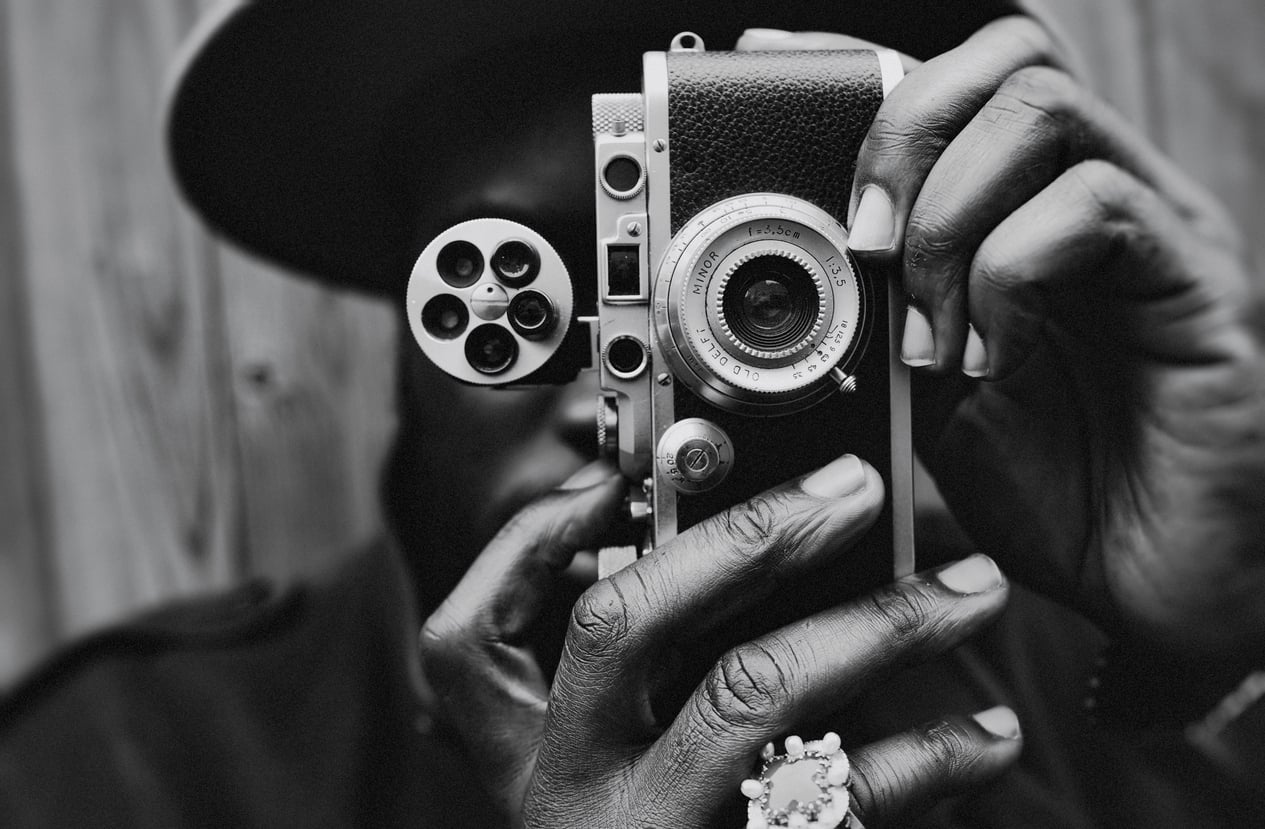 A person holding an old-fashioned movie camera close to their face, focusing through the viewfinder—a nostalgic reminder of life before smartphones. The image is in black and white, with a blurred wooden background.
