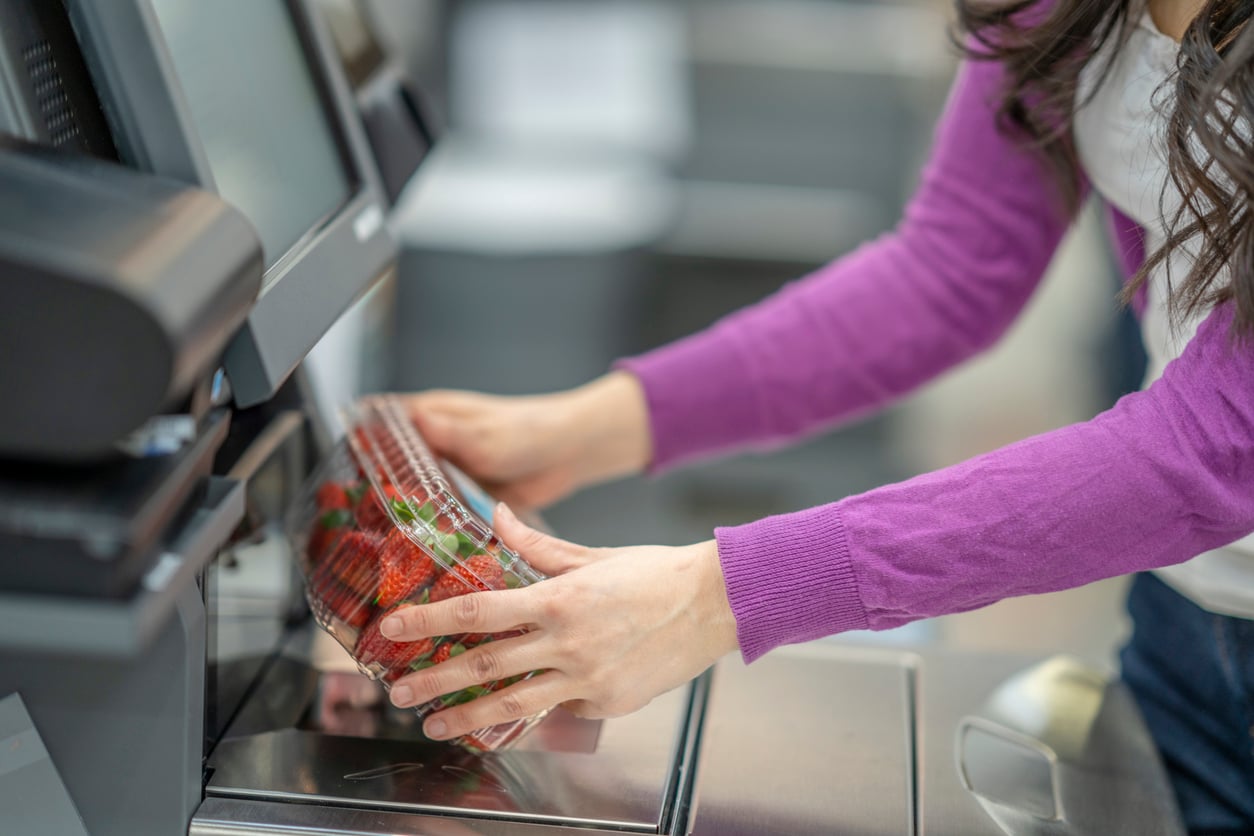A person in a purple sweater scans a plastic container of strawberries at a self-checkout station in a grocery store.