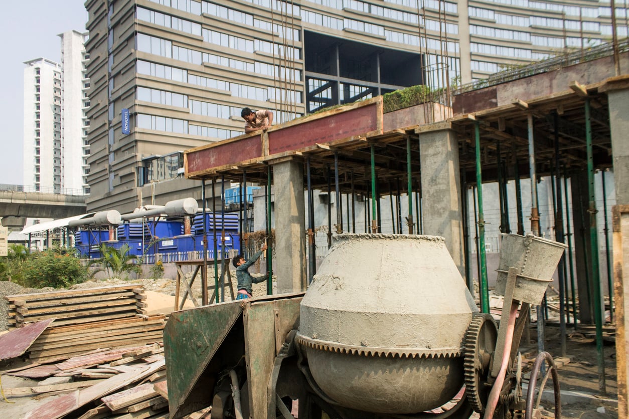 A construction site with workers on scaffolding, a concrete mixer, stacked building materials, and tall modern buildings in the background under a clear sky—much like avoiding hotel booking mistakes, attention to detail ensures every element comes together seamlessly.
