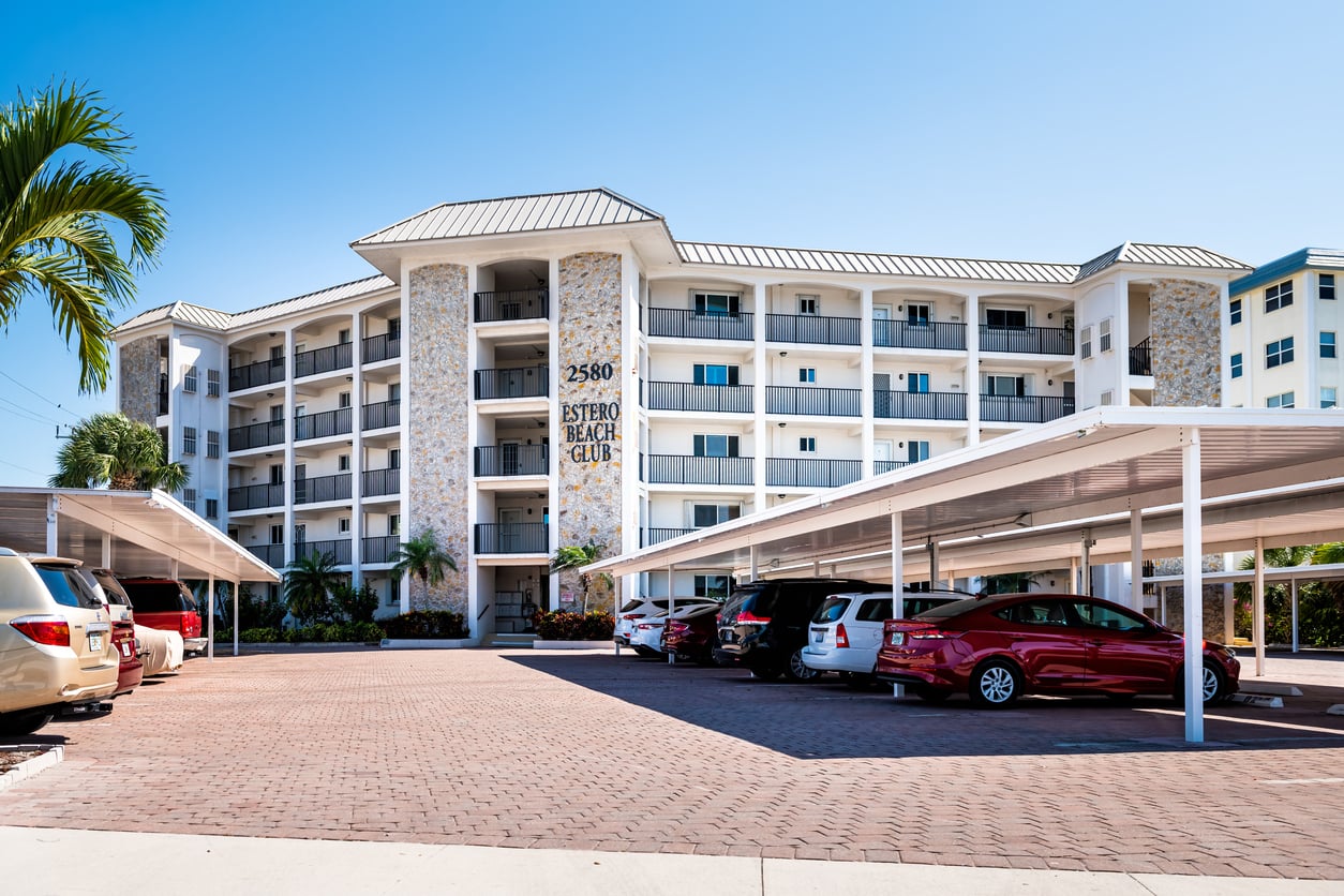 A four-story condominium building with stone accents and balconies sits behind a brick-paved parking lot filled with cars. A sign reads "2580 Estero Beach Club," offering beach vacation savings. Palm trees and clear blue sky are visible.