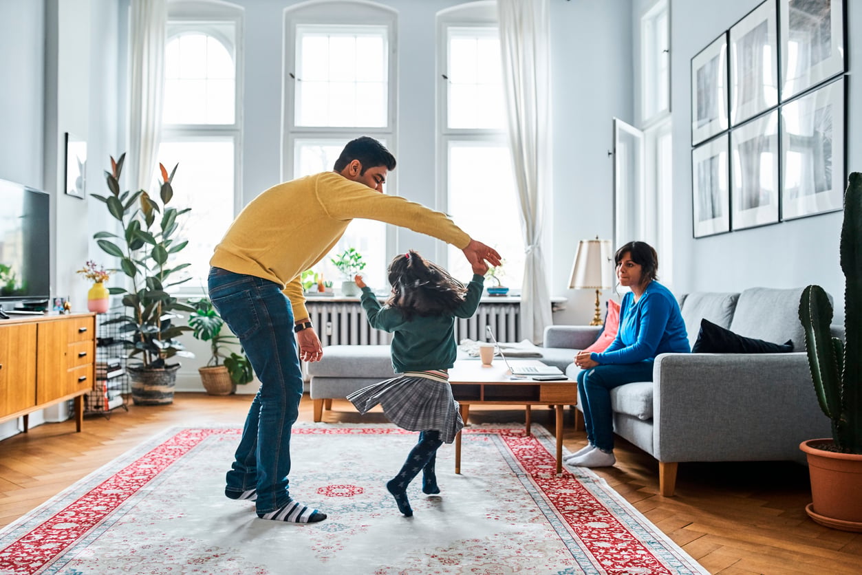 A man twirls a young girl in a bright living room while a woman sits on a gray couch, watching them—a joyful moment, even as families in America are having fewer children. The room features large windows, wooden furniture, plants, and a red-patterned rug.