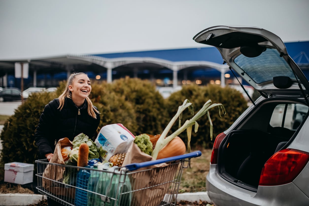 A smiling woman stands outdoors beside a car, loading groceries from a full shopping cart into the open trunk, managing household expenses in a busy parking lot.