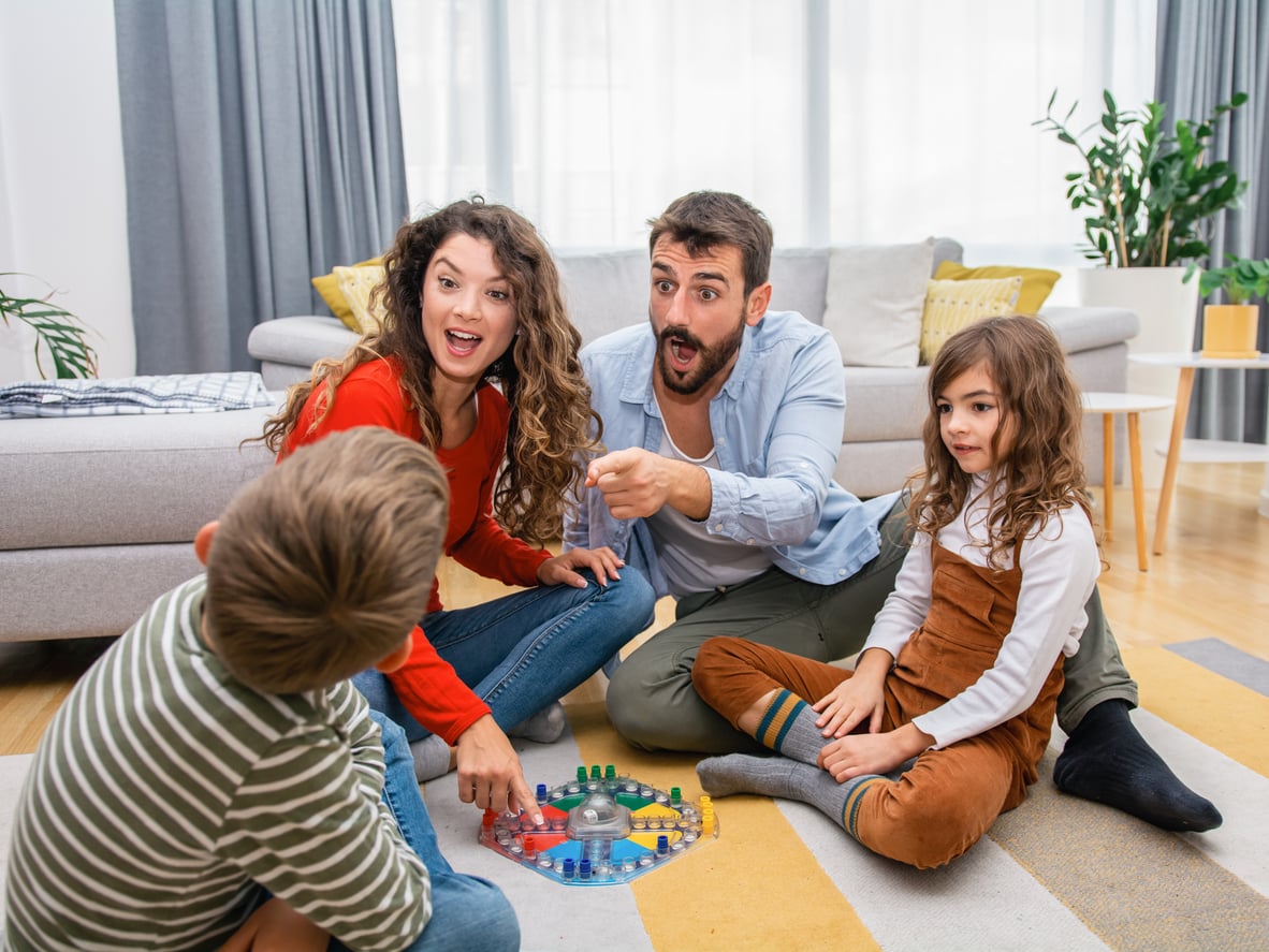 A family of four sits on the floor playing a board game in a living room. The parents look excited, and the two children are engaged in the game, with colorful pieces on the board between them—a perfect example of money saving habits as they enjoy quality time together without spending much.