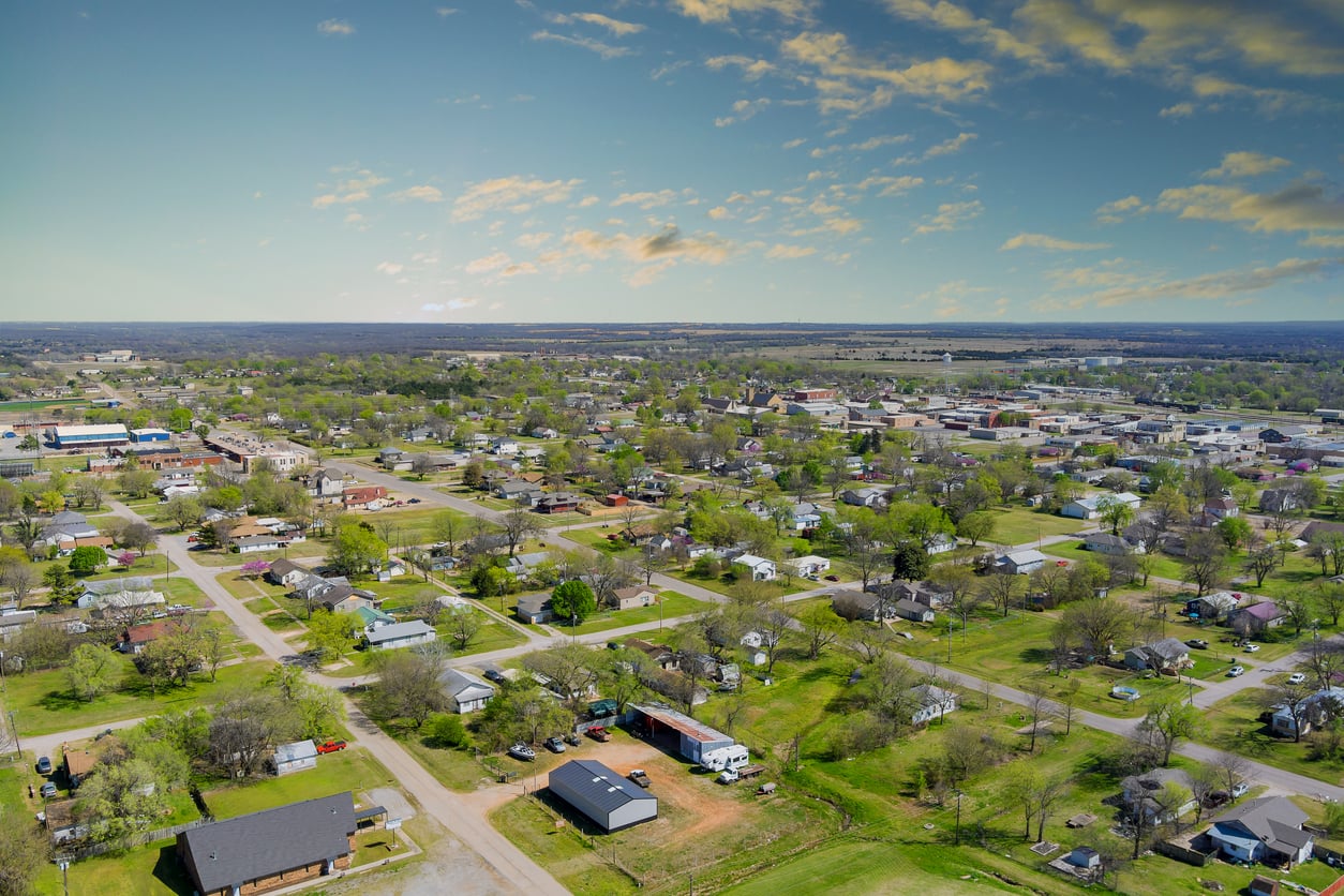 Aerial view of a suburban neighborhood with houses, streets, and green lawns under a partly cloudy sky, extending toward a distant horizon—capturing the quintessential charm of American towns.