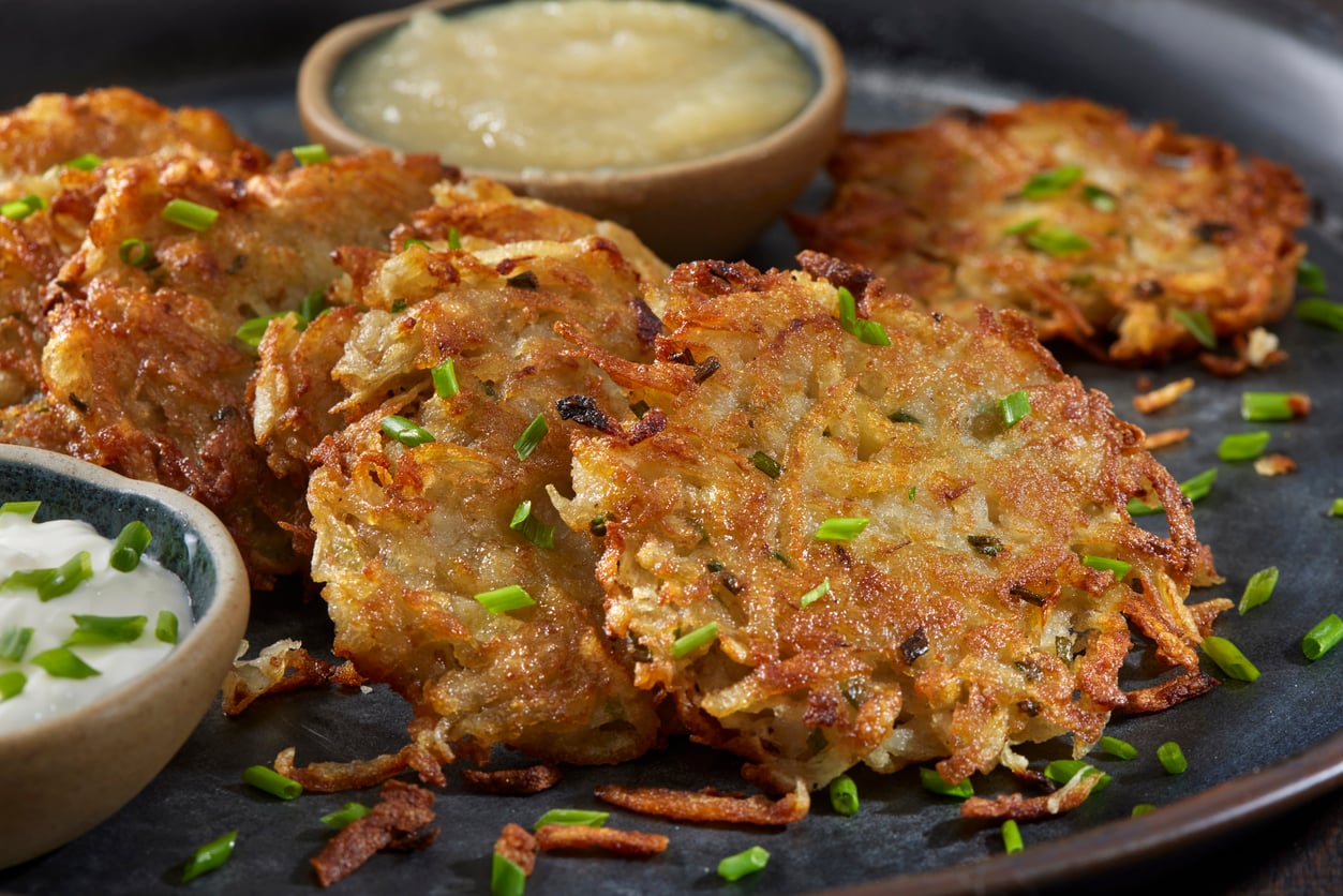 Crispy potato latkes garnished with chopped chives, served on a dark plate with small bowls of applesauce and sour cream on the side.