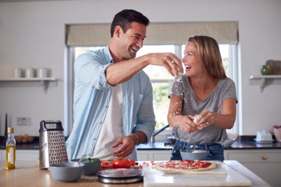 A man and woman laugh while making pizza together in a bright kitchen, with best shredded cheese, tomatoes, and olive oil on the counter.