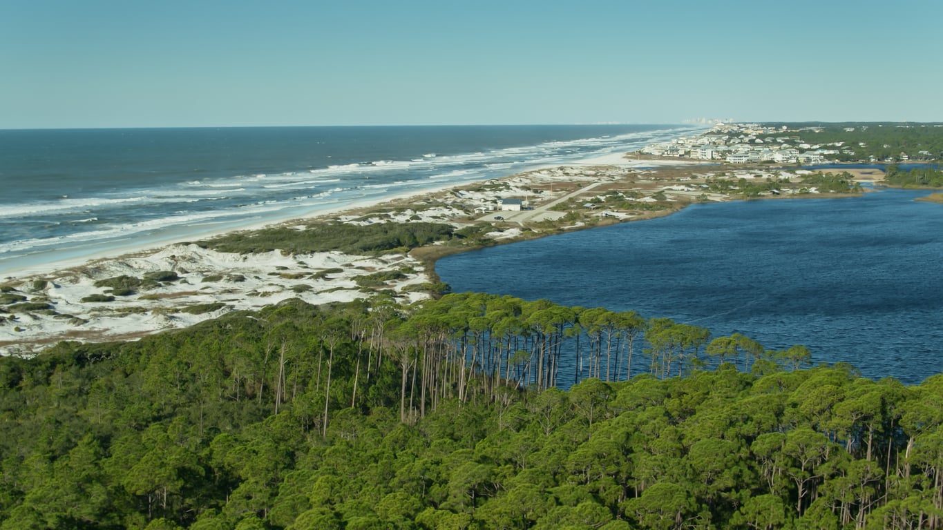 Aerial view of a coastal landscape with green forest, a blue lake, white sand dunes, and waves along the shoreline, with beach towns and buildings visible in the distance under a clear sky.