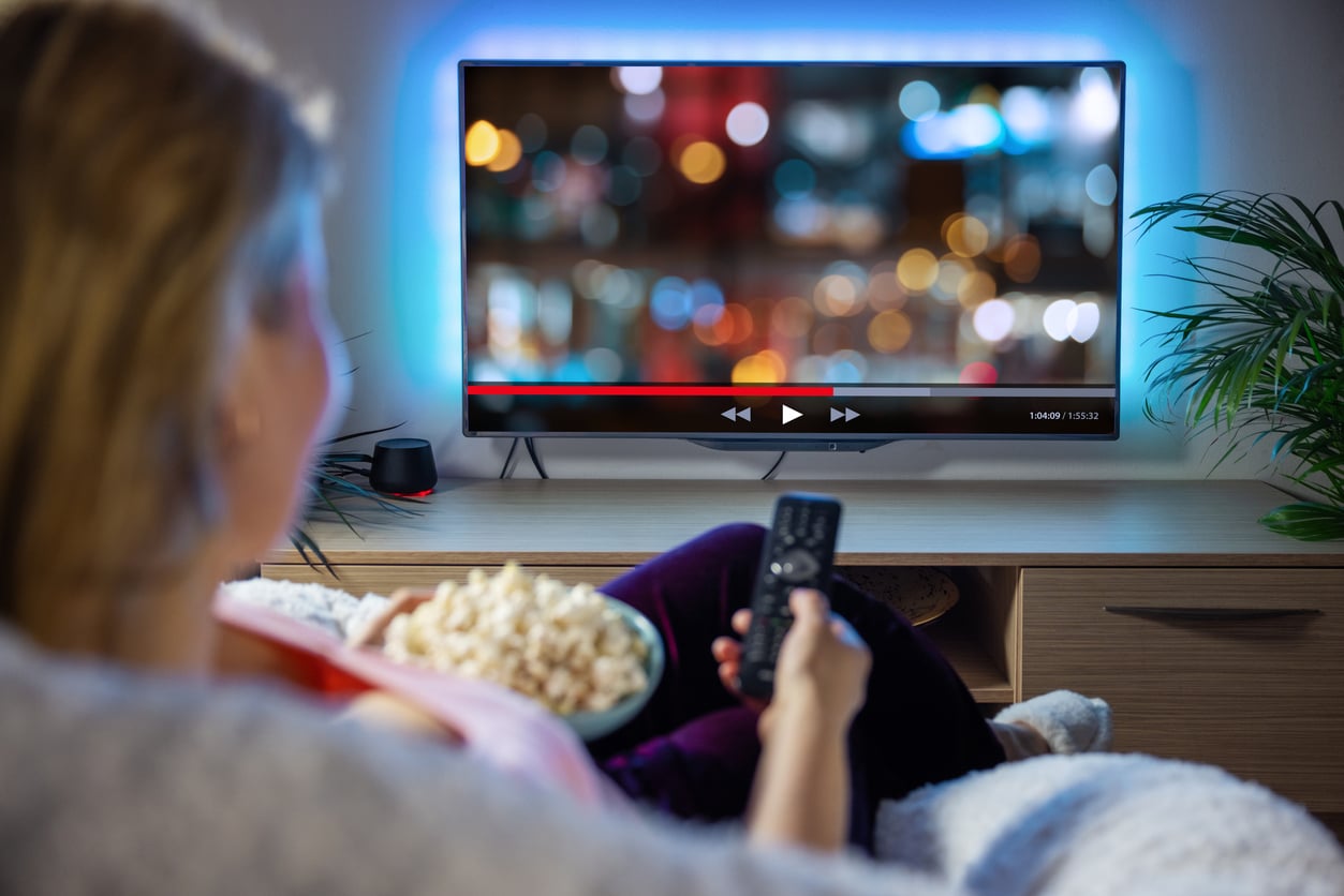 A person holding a remote and a bowl of popcorn sits on a couch watching a streaming video with a blurred screen on a wall-mounted TV in a cozy living room.