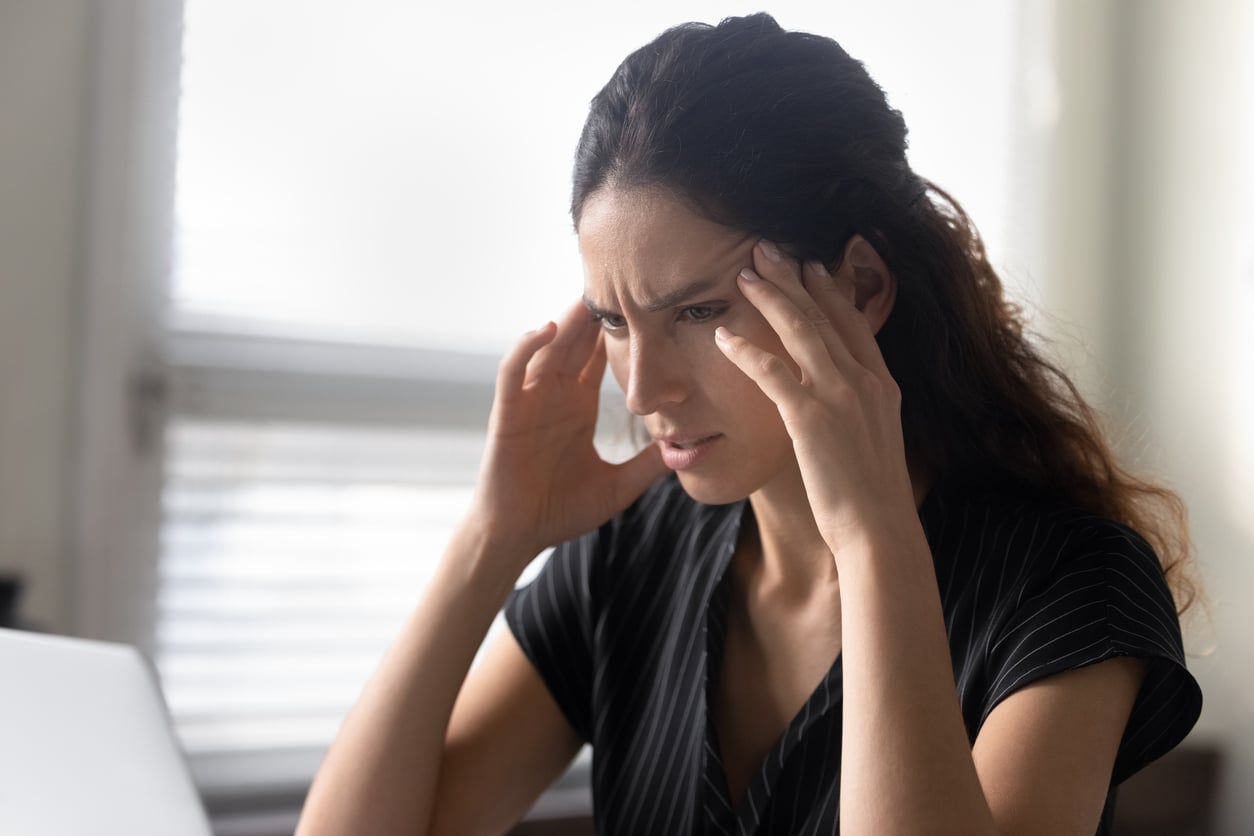 A woman sits at a desk, touching her temples and looking stressed or worried while staring at a laptop screen, perhaps longing for the simplicity of life before smartphones.