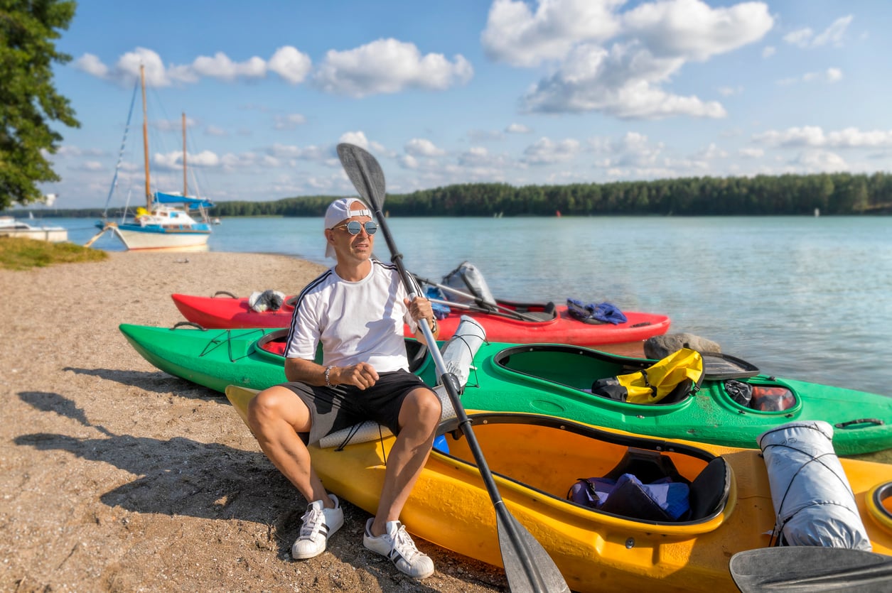 A man in a white t-shirt and cap sits on a yellow kayak during his beach vacation, holding a paddle. Colorful kayaks and gear surround him, with a lake, sailboats, and trees in the background under a partly cloudy sky.