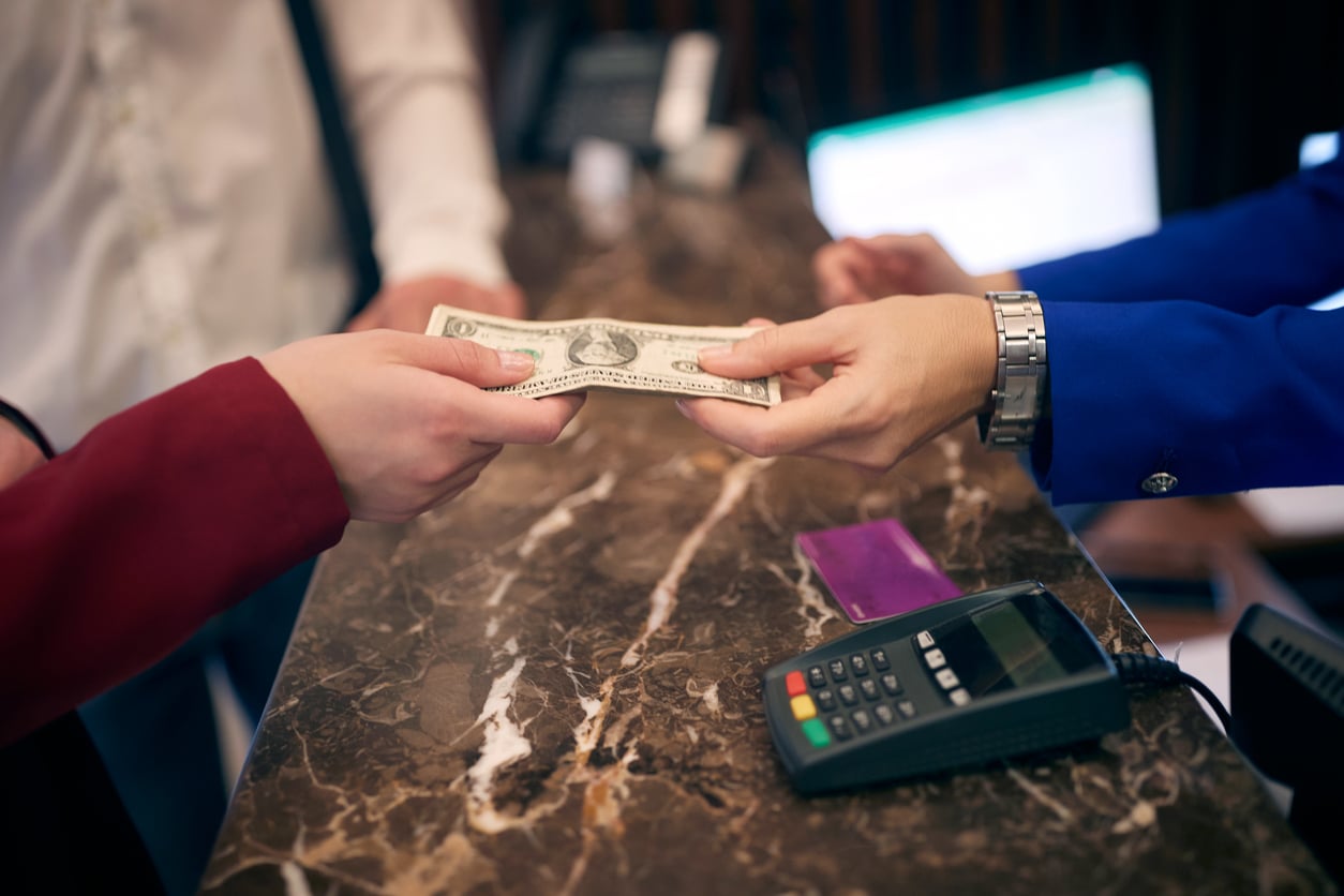 Two people exchange a US dollar bill across a marble counter, with a credit card, a payment terminal, and a computer keyboard nearby—planning beach vacation savings for their next getaway.