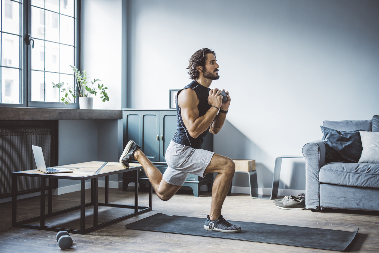 A man in athletic wear performs a Bulgarian split squat on a yoga mat in his living room, holding a dumbbell at his chest. Exercising at home with simple gear like a laptop and dumbbell is a great way to save money while staying fit.