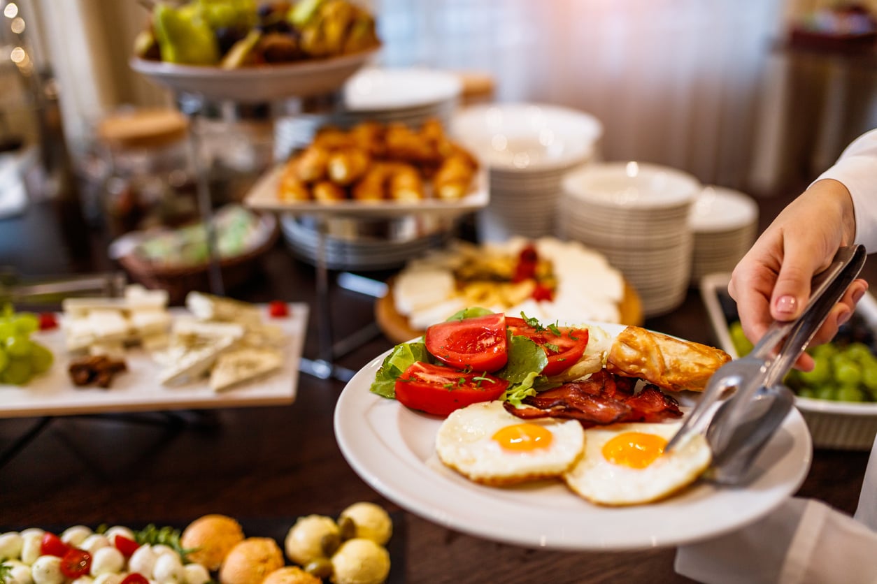 A hand uses tongs to serve breakfast onto a plate with sunny-side-up eggs, tomato slices, bacon, and a pastry, with assorted breakfast foods and cheeses in the background.