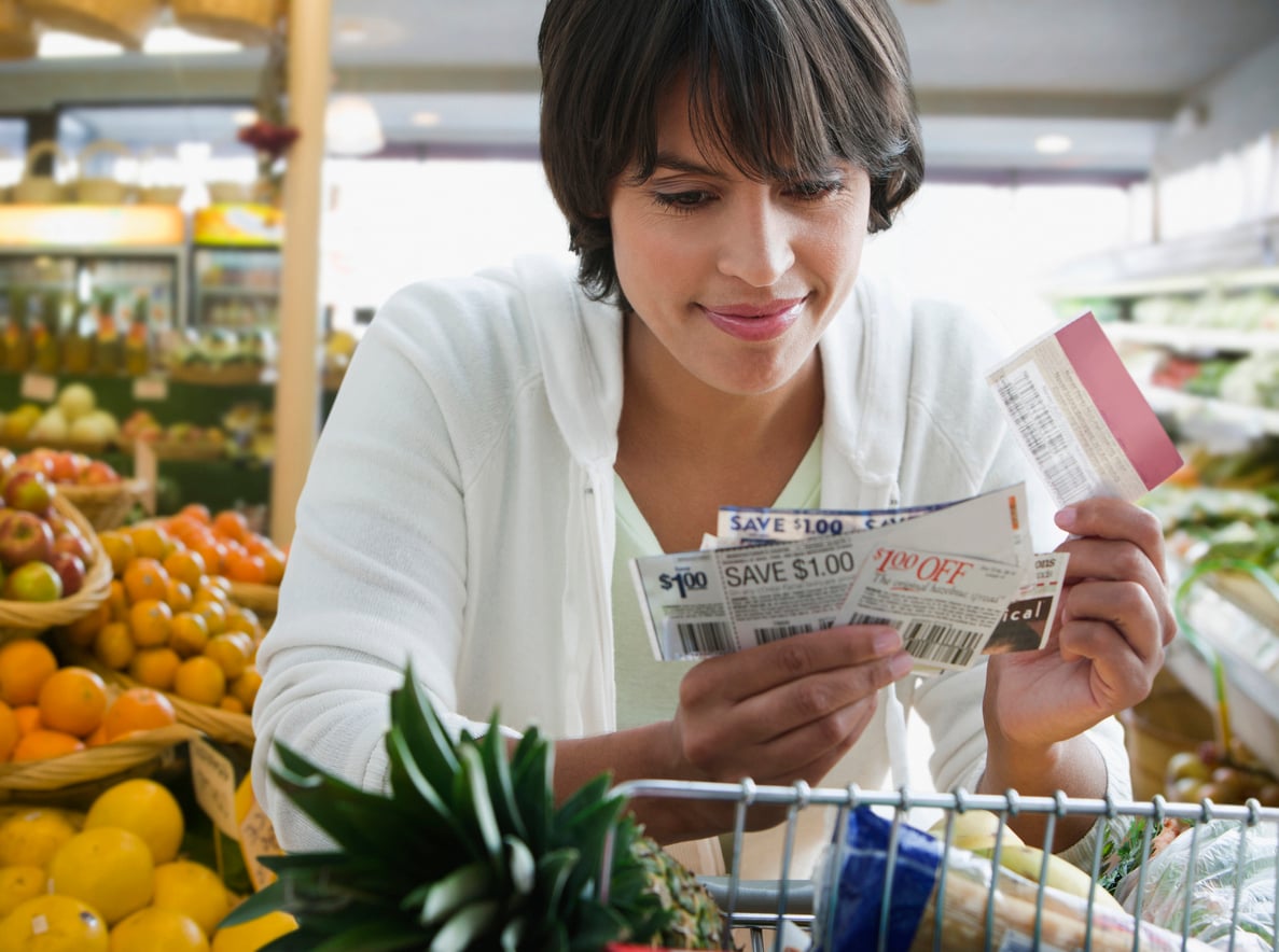 A woman in a grocery store looks at several coupons while standing behind a shopping cart filled with fruits and vegetables.