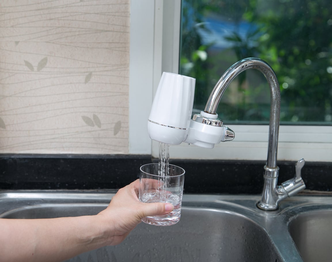 A hand holds a glass under a kitchen sink faucet with a white water filter attached, filling the glass with water—an easy way to reduce household expenses. The sink and faucet are in front of a window showing greenery outside.