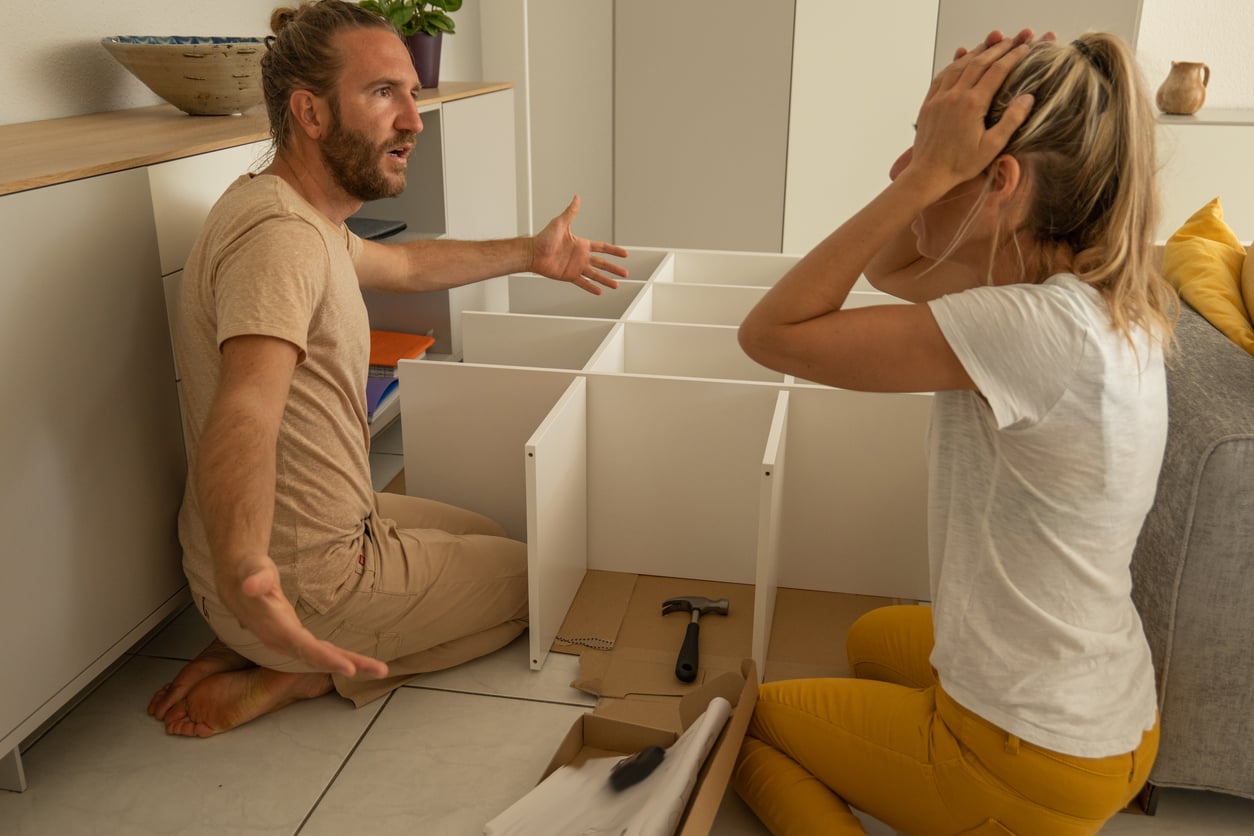 A man and woman sit on the floor assembling furniture. The man looks frustrated with arms outstretched, while the woman holds her head in her hands, surrounded by tools and unassembled parts—clearly fed up with products that don't last.
