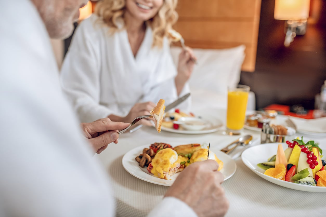 Two people in white robes enjoy a breakfast in bed with eggs, fruit, and juice—an idyllic morning that highlights the perks of a well-planned stay. The focus is on the beautifully plated food, with their faces partially out of frame and blurred in the background—a scene made perfect by avoiding common hotel booking mistakes.
