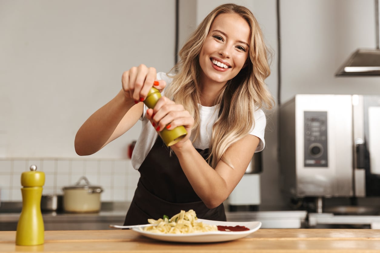 Smiling woman in an apron grinds pepper onto a plate of pasta in a kitchen. A pepper grinder and appliances are visible in the background.