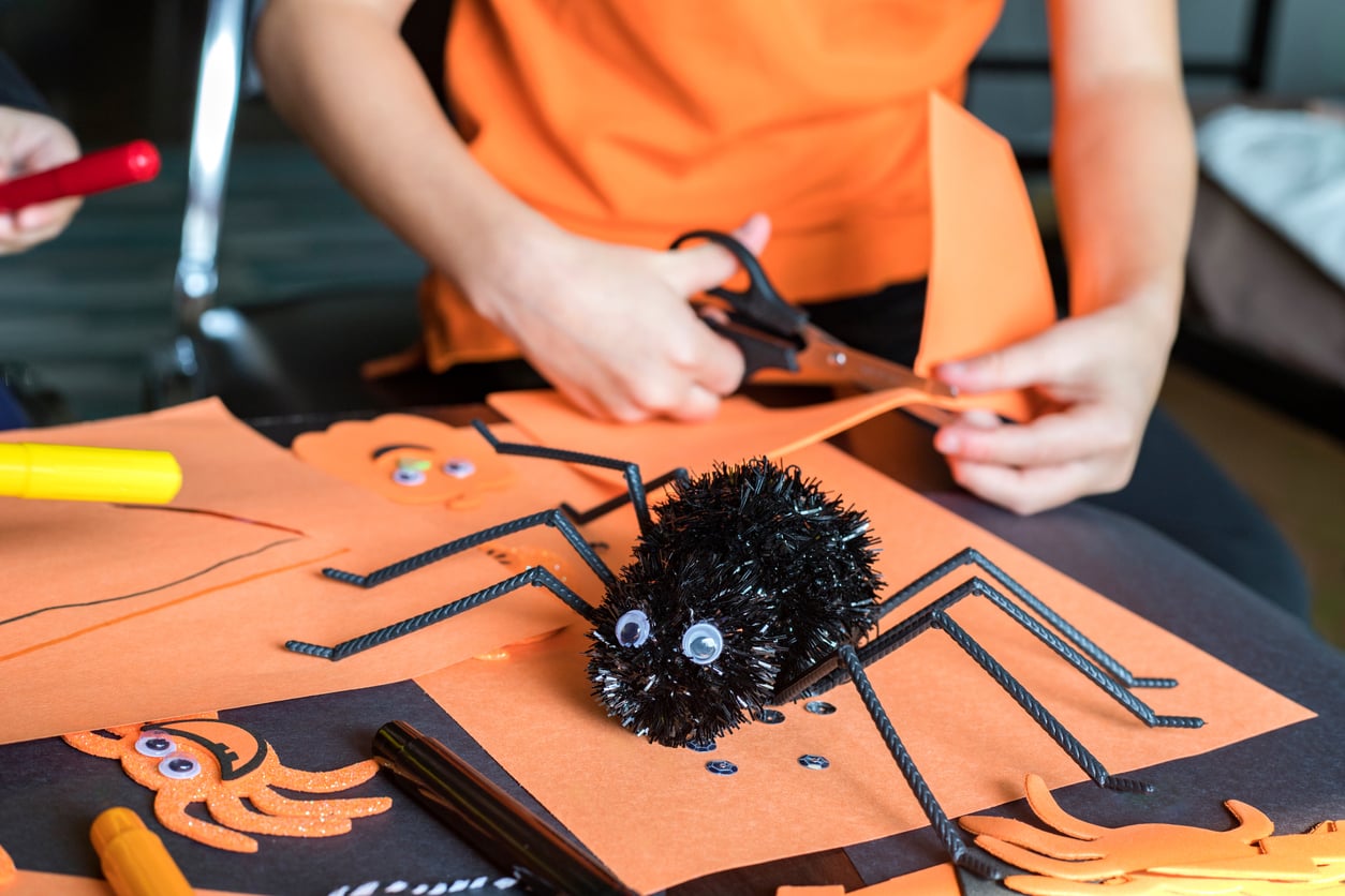 A person cuts orange paper for a Halloween craft—one of many fun Halloween activities. A black spider decoration with googly eyes and pipe cleaner legs sits on the table with markers and other orange cutouts.