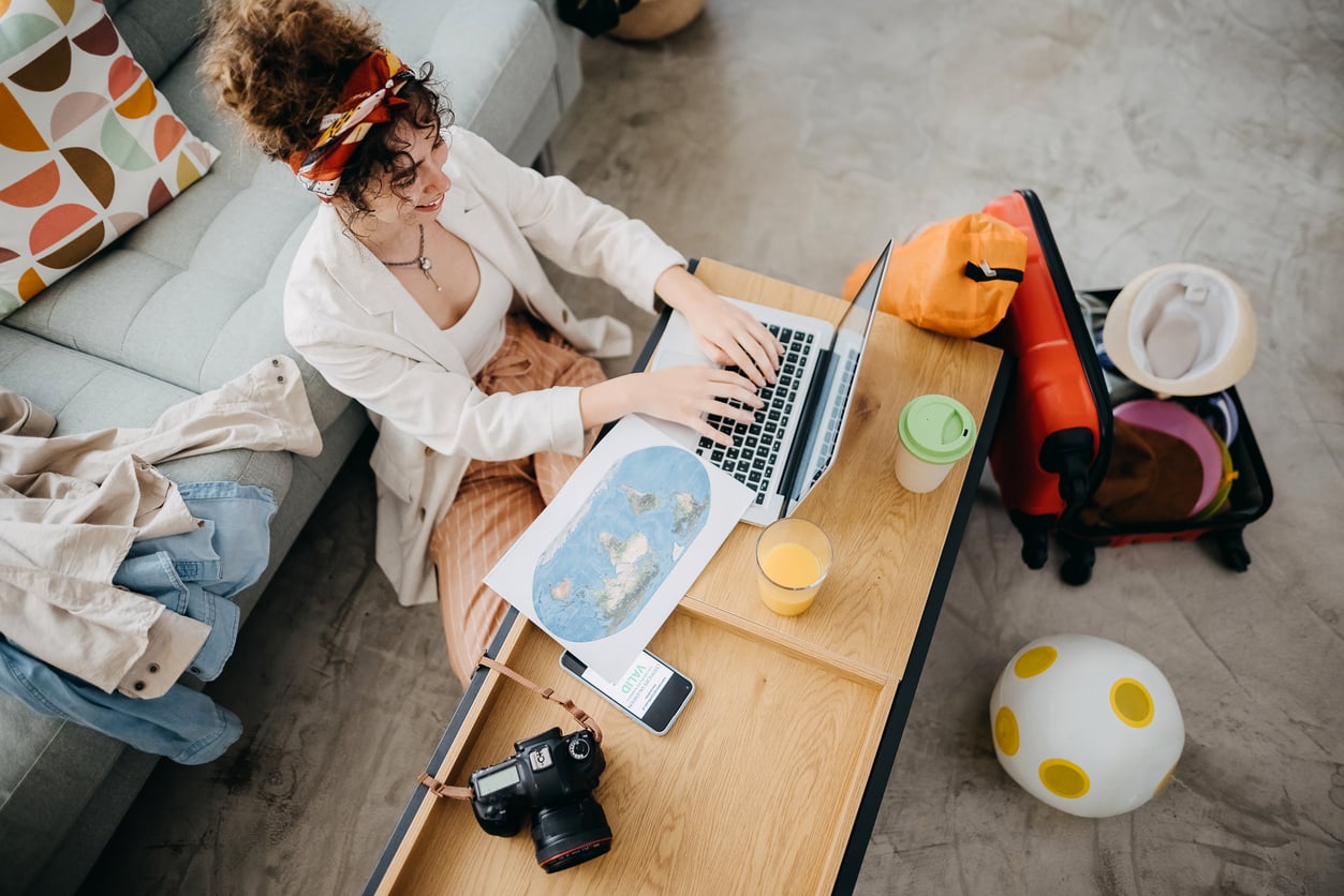 A person sits at a table using a laptop, with a map, camera, phone, drink, and juice nearby. As women are changing their spending behaviors, the scene hints at thoughtful planning and mindful choices—suggested by the organized setup. A suitcase and toy ball are on the floor; a couch with colorful pillows is in the background.
