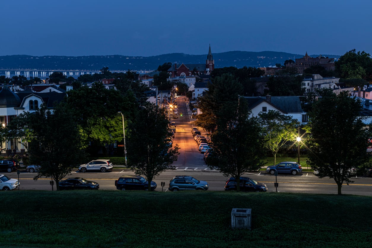 Evening view of a small city street lined with houses and parked cars, illuminated by streetlights, evokes the cozy charm found in some of the best trick or treat towns in America, with hills and a bridge visible in the background.