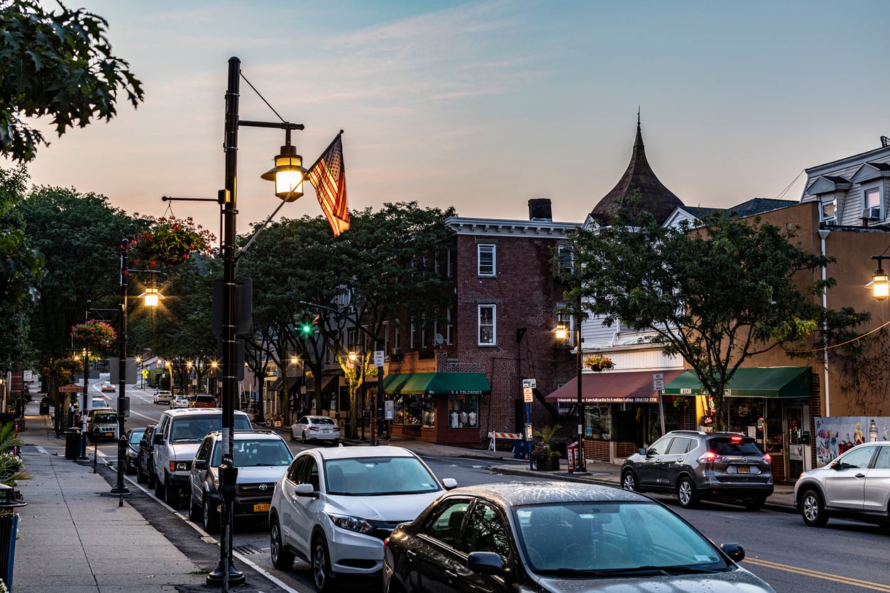 Cars parked along a quiet small-town street at dusk, with glowing streetlights casting a gentle glow on Halloween decorations, an American flag, shops, and trees lining the sidewalk. A building with a pointed roof is visible in the background.