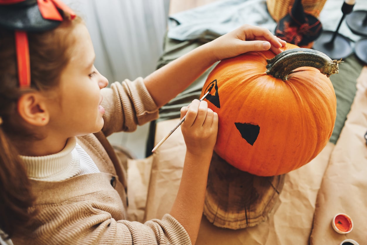 A child paints black triangle eyes on a pumpkin, preparing a jack-o'-lantern for Halloween. She is focused on her art, with supplies and decorations visible on the table—one of many fun Halloween activities enjoyed this season.