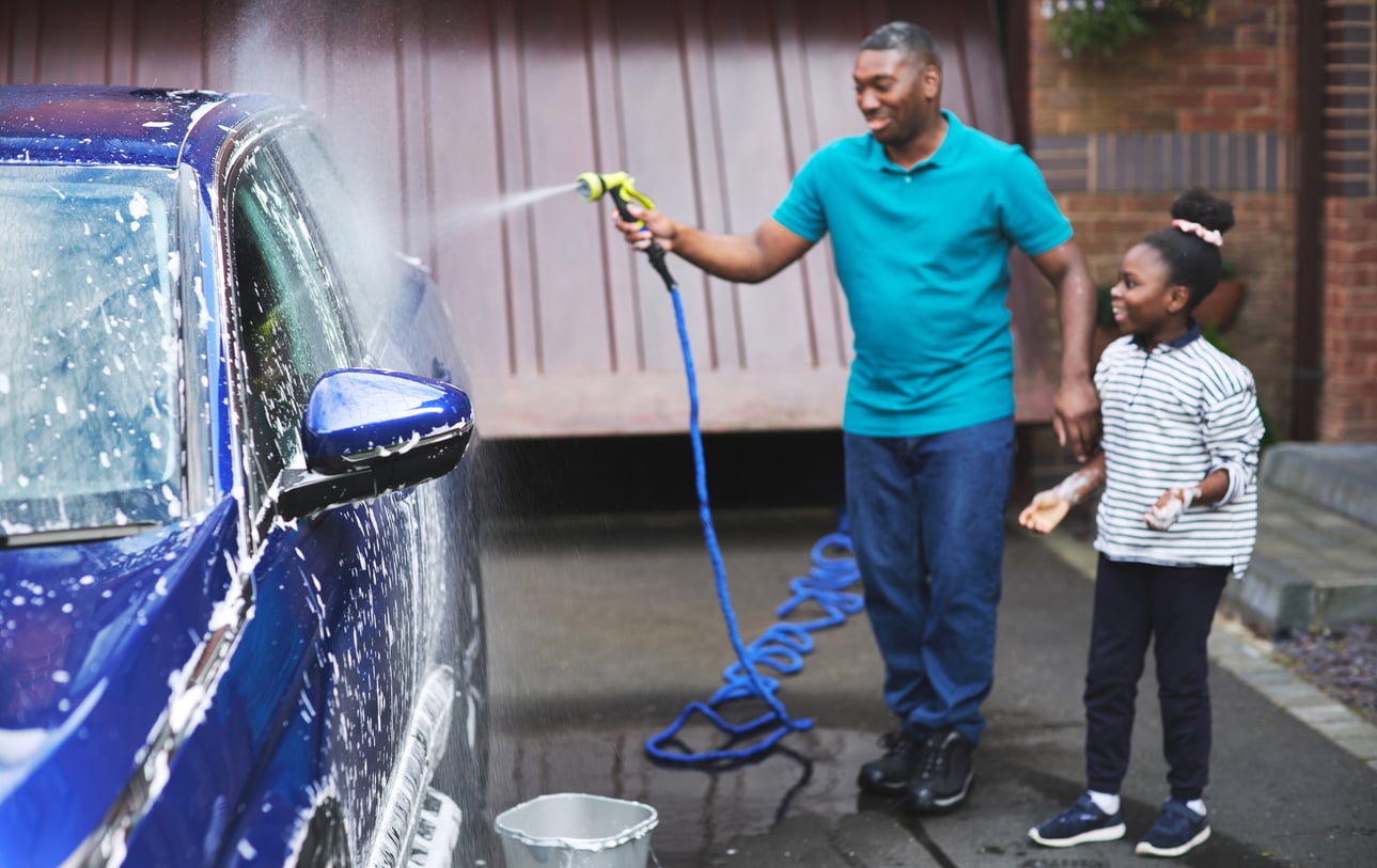 A man and a young girl are washing a blue car together outdoors. The man is spraying water on the car while the girl stands beside him, smiling. Soap suds cover the car, and a bucket is on the ground nearby.