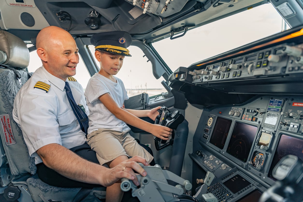 A pilot smiles while sitting beside a young boy wearing a captain’s hat, who is holding the controls in an airplane cockpit filled with various instruments and displays.