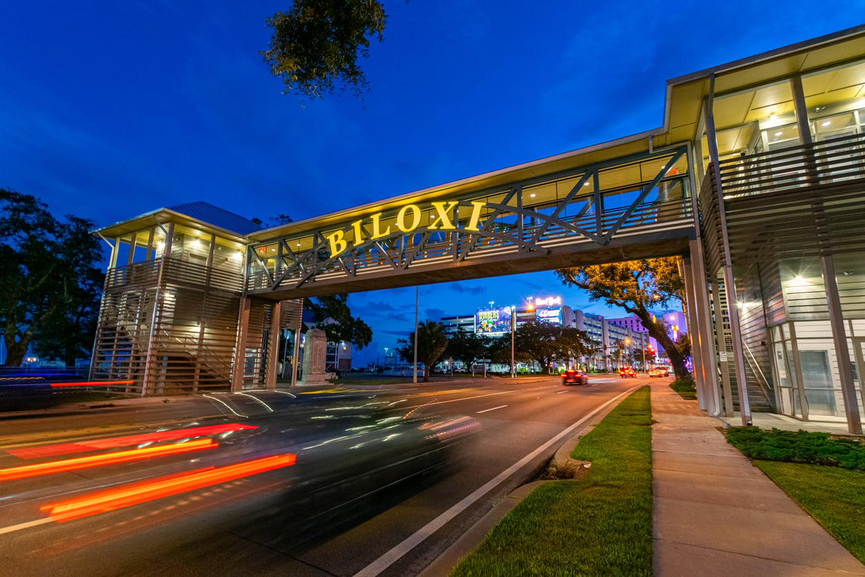 A pedestrian bridge with "BILOXI" in bright yellow letters spans a busy street at dusk, with blurred car lights and nearby buildings illuminated—an inviting scene in one of the Gulf Coast’s most affordable travel destinations.