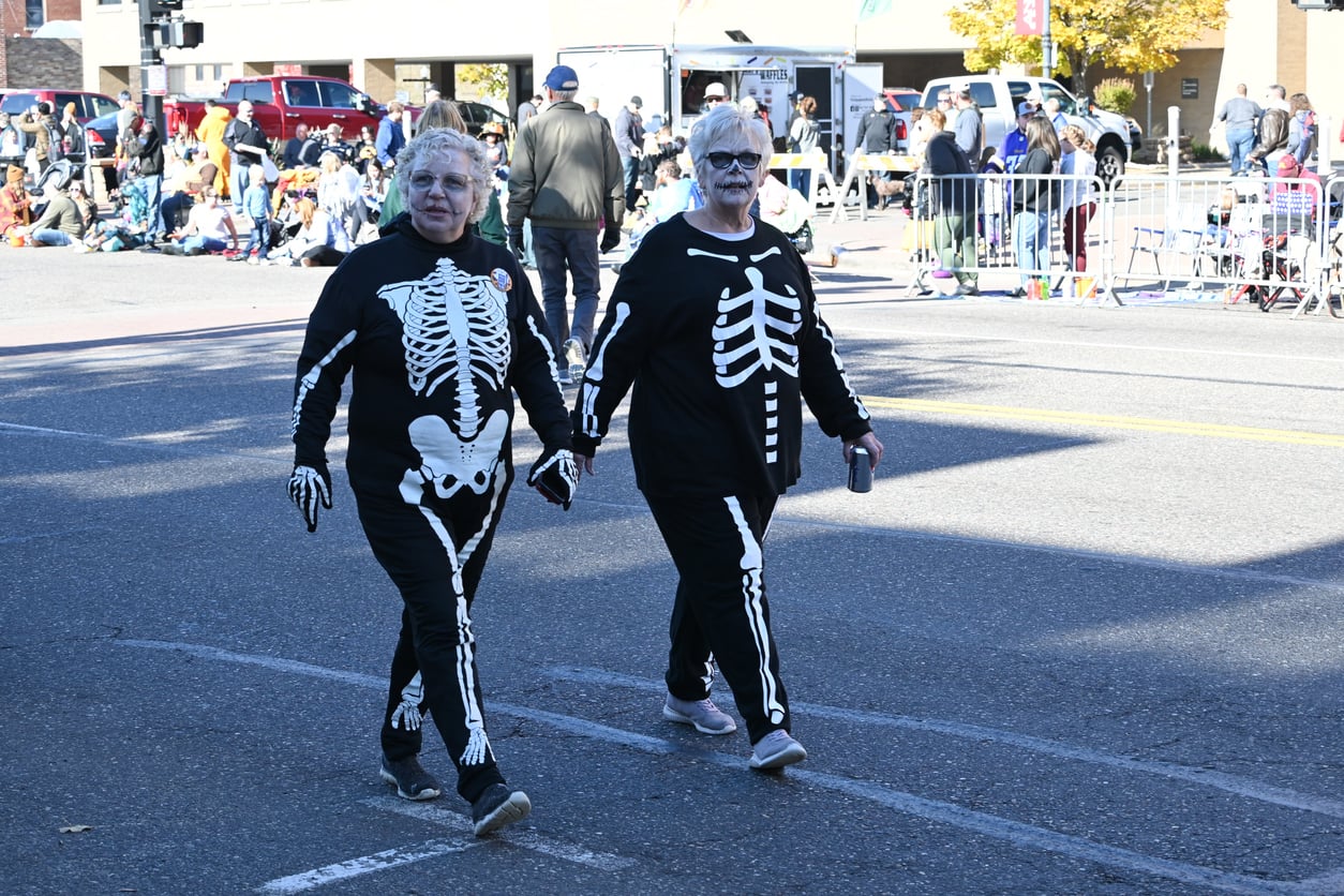 Two people wearing Halloween skeleton costumes and makeup walk hand in hand down a street during a parade, with a crowd of spectators in the background.