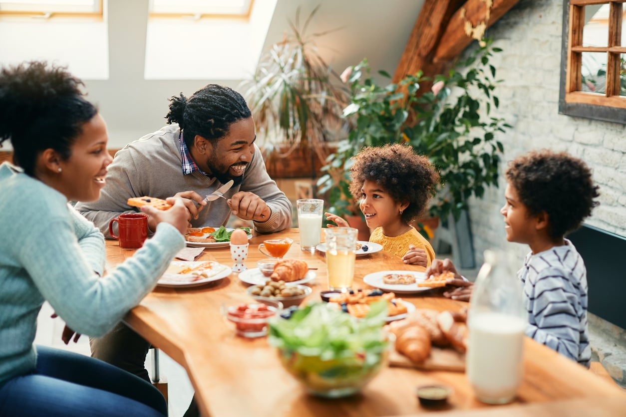 A family of four enjoys breakfast together at a wooden table, smiling and laughing, reminiscent of life before smartphones. The table is filled with food including salad, croissants, and juice as sunlight streams in from the windows above.