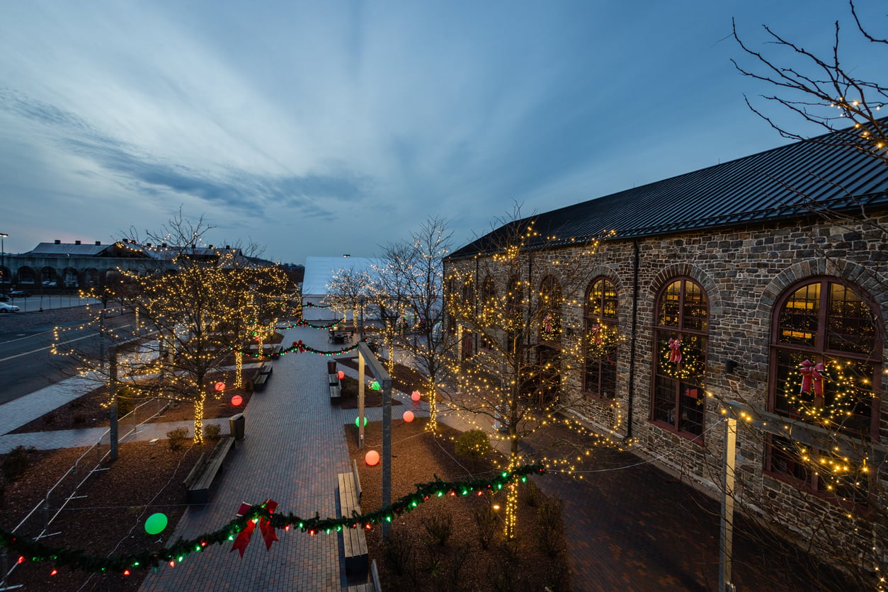 A brick walkway lined with trees decorated in yellow holiday lights, colorful ornaments, and garlands, beside a large stone building with wreaths on the windows at dusk—capturing the festive spirit found in the best Christmas towns in America.
