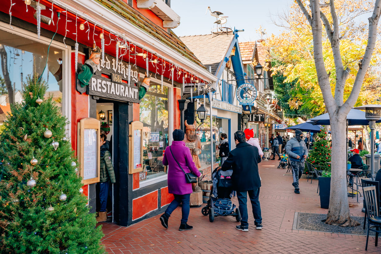 People walk along a charming street with shops and a restaurant; holiday decorations and Christmas trees are visible, capturing the festive spirit that makes this one of the best Christmas towns in America. Some people are seated outdoors at a café, enjoying the cheerful atmosphere.