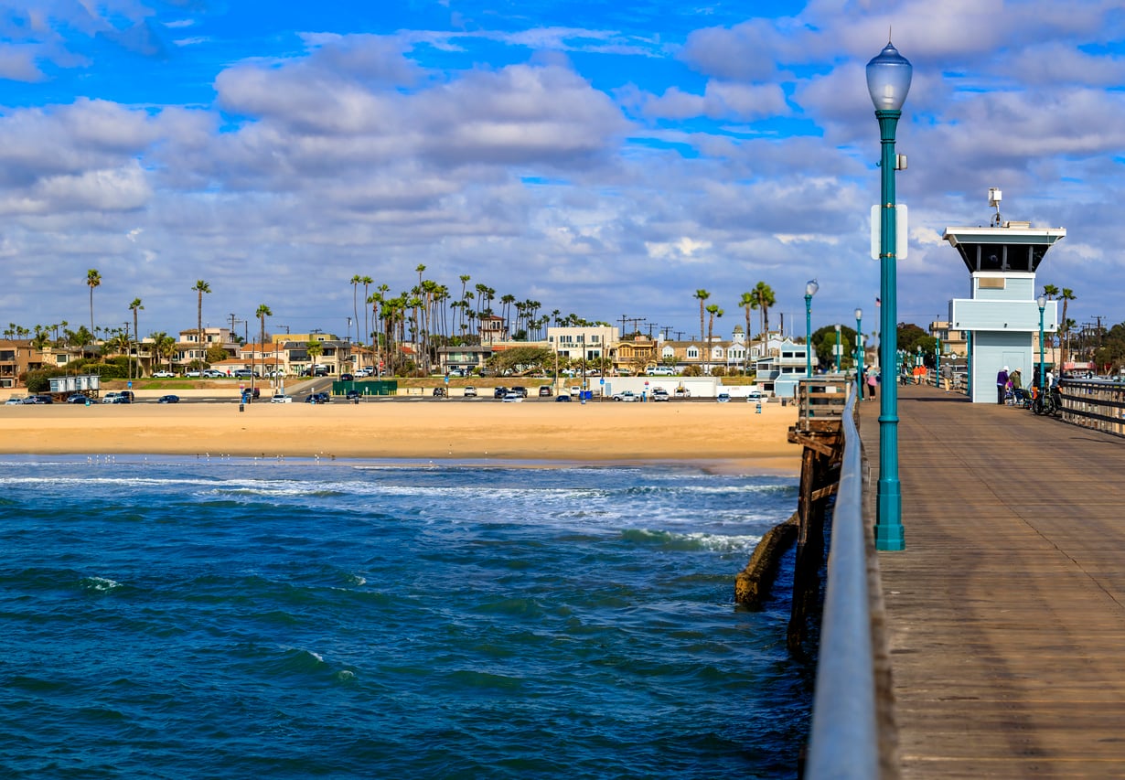 A wooden pier extends over blue ocean water toward a sandy beach lined with palm trees and houses, evoking the charm of classic beach towns under a partly cloudy sky.