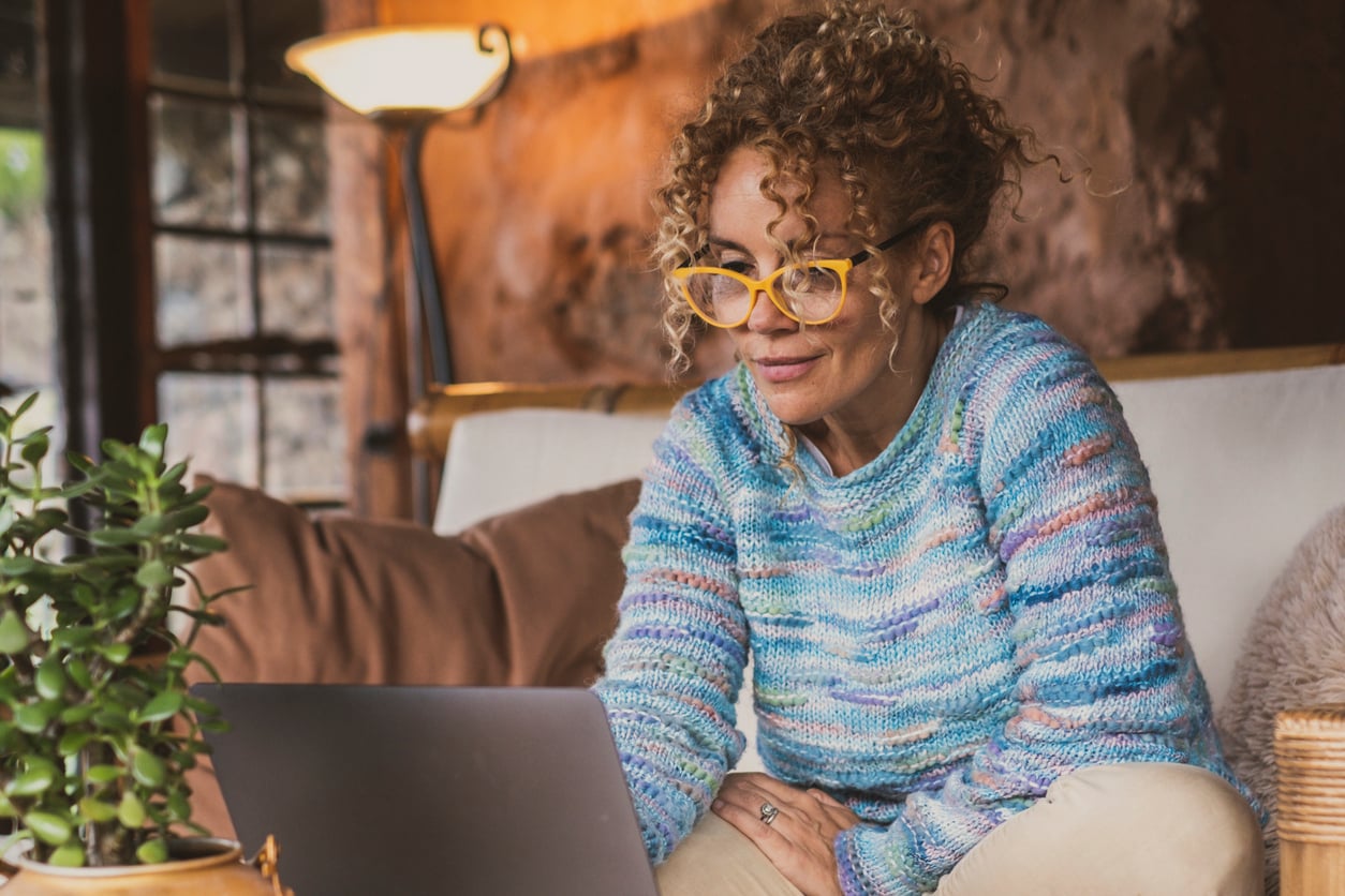 A woman with curly hair and yellow glasses sits on a couch, smiling while looking at a laptop. She wears a colorful sweater and is in a cozy, warmly lit room with a plant and lamp nearby—perhaps double-checking her travel plans to avoid common hotel booking mistakes.
