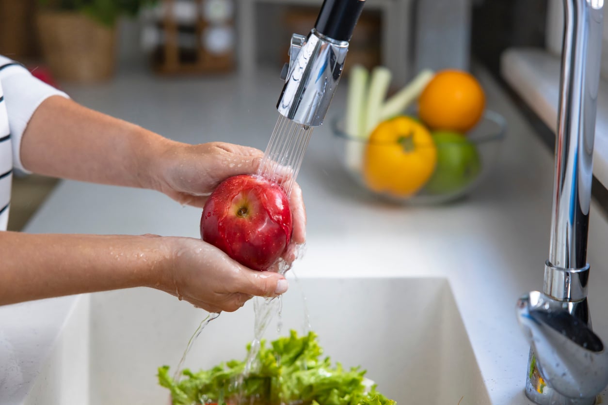 A person rinses a red apple under running water in a kitchen sink filled with green lettuce. In the background, a glass bowl with yellow bell peppers and other vegetables sits on the counter.