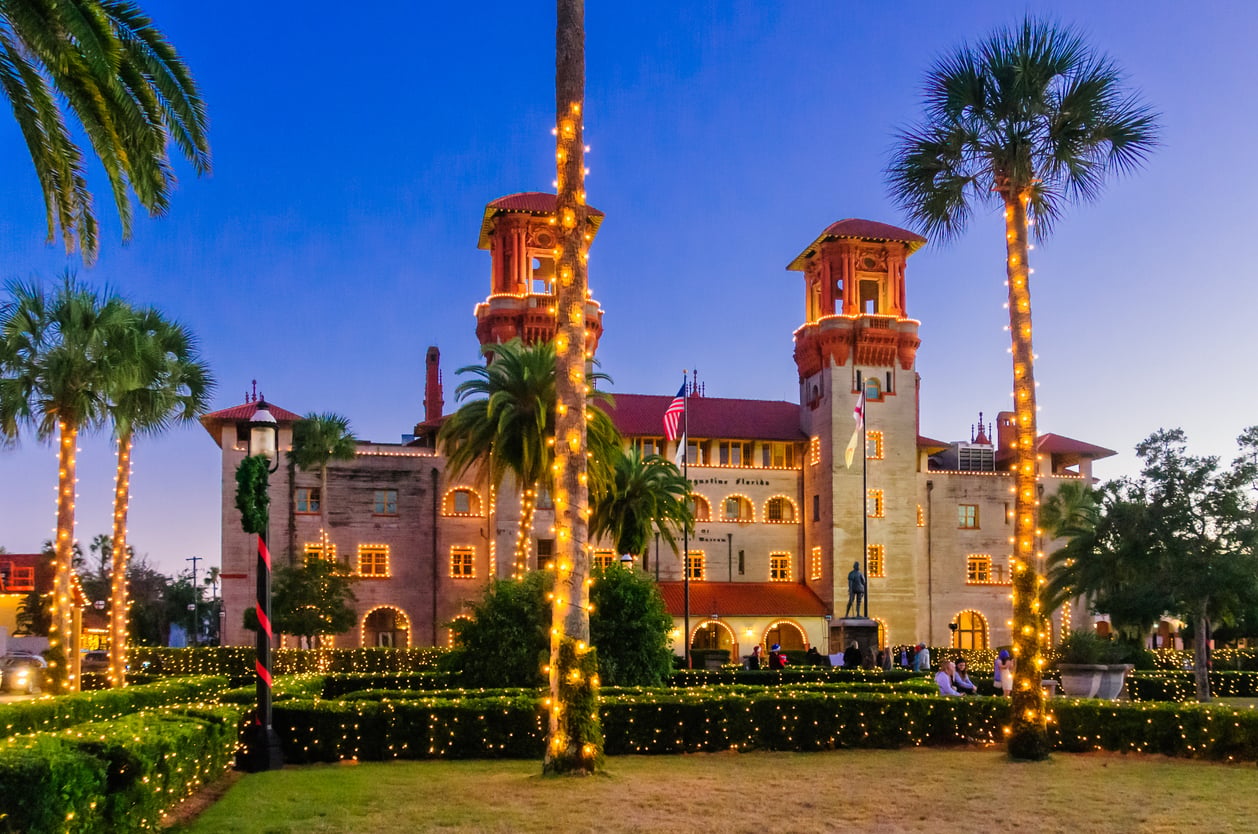 A historic building with two towers is adorned with festive lights, capturing the charm often found in the best Christmas towns in America. Palm trees wrapped in string lights frame the scene at dusk, and the sky is clear and blue.
