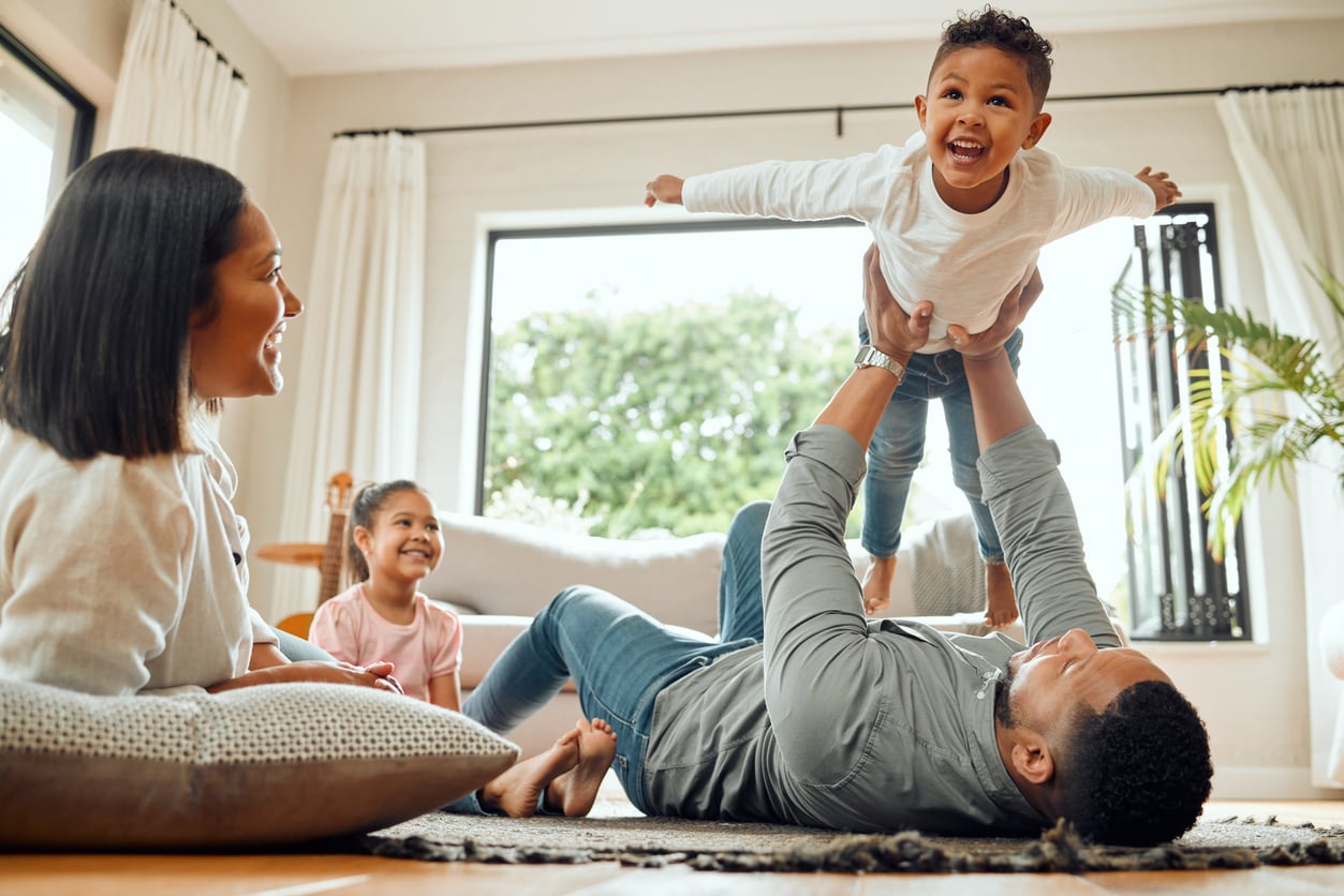 A family of four enjoys time together indoors, making the most of the benefits of renting. A man lies on his back, lifting a smiling young boy in the air as a woman and girl sit nearby, laughing under sunlight streaming through large windows.