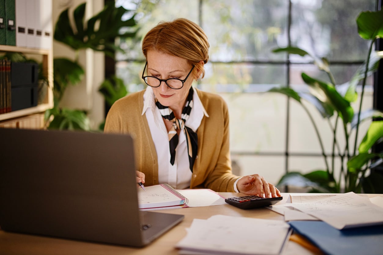 A woman with glasses works at a desk with a laptop, calculator, and papers, surrounded by green plants and sunlight coming through a large window—a scene that quietly reflects how women are changing their spending behaviors in thoughtful, intentional ways.