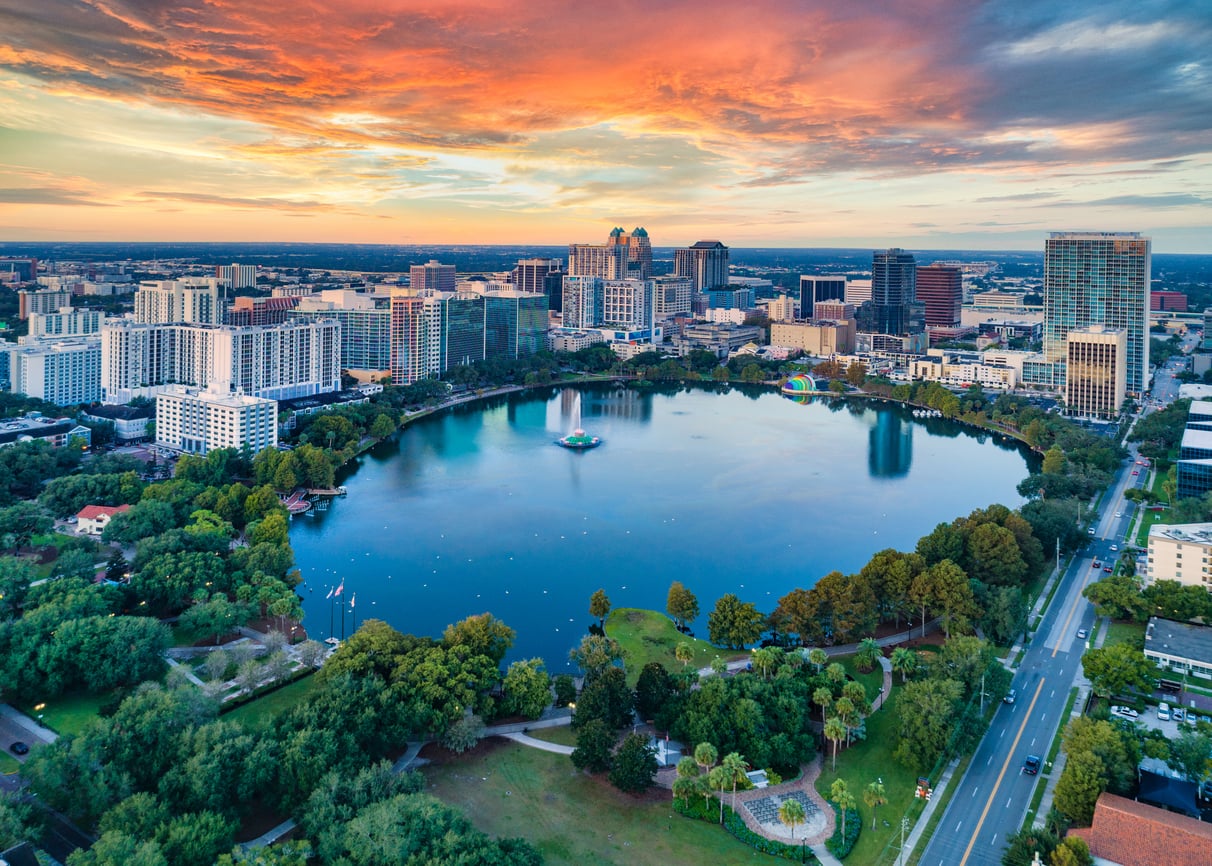 Aerial view of a travel-friendly city skyline at sunset with a large round lake surrounded by trees and buildings, vibrant sky with orange and blue hues.