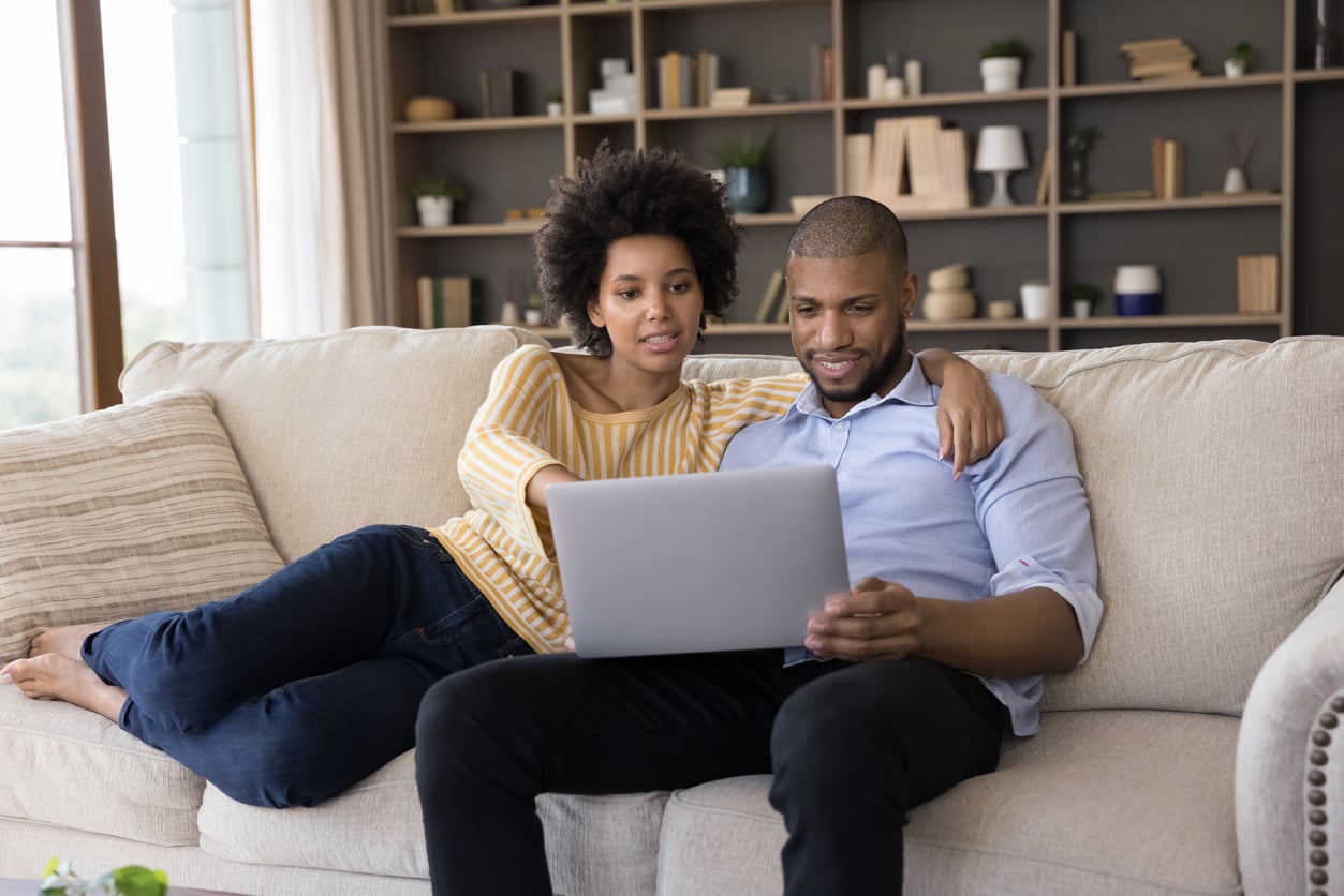 A couple sits on a sofa at home, looking at a laptop together as they discuss hotel booking tips. The woman points at the screen while the man holds the laptop on his lap. Shelves with books and decor fill the background.