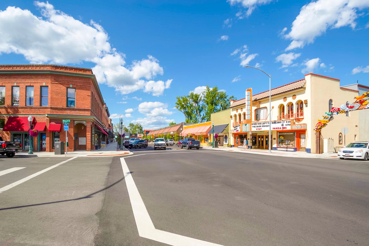 A sunny street scene with historic brick buildings, colorful awnings, parked cars, and a theater with a marquee. The sky is blue with scattered clouds.