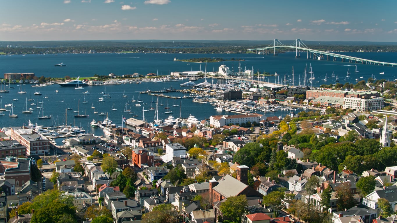 Aerial view of a travel-friendly coastal city with a marina full of boats, a large bridge over blue water, and numerous houses and trees in the foreground under a clear sky.