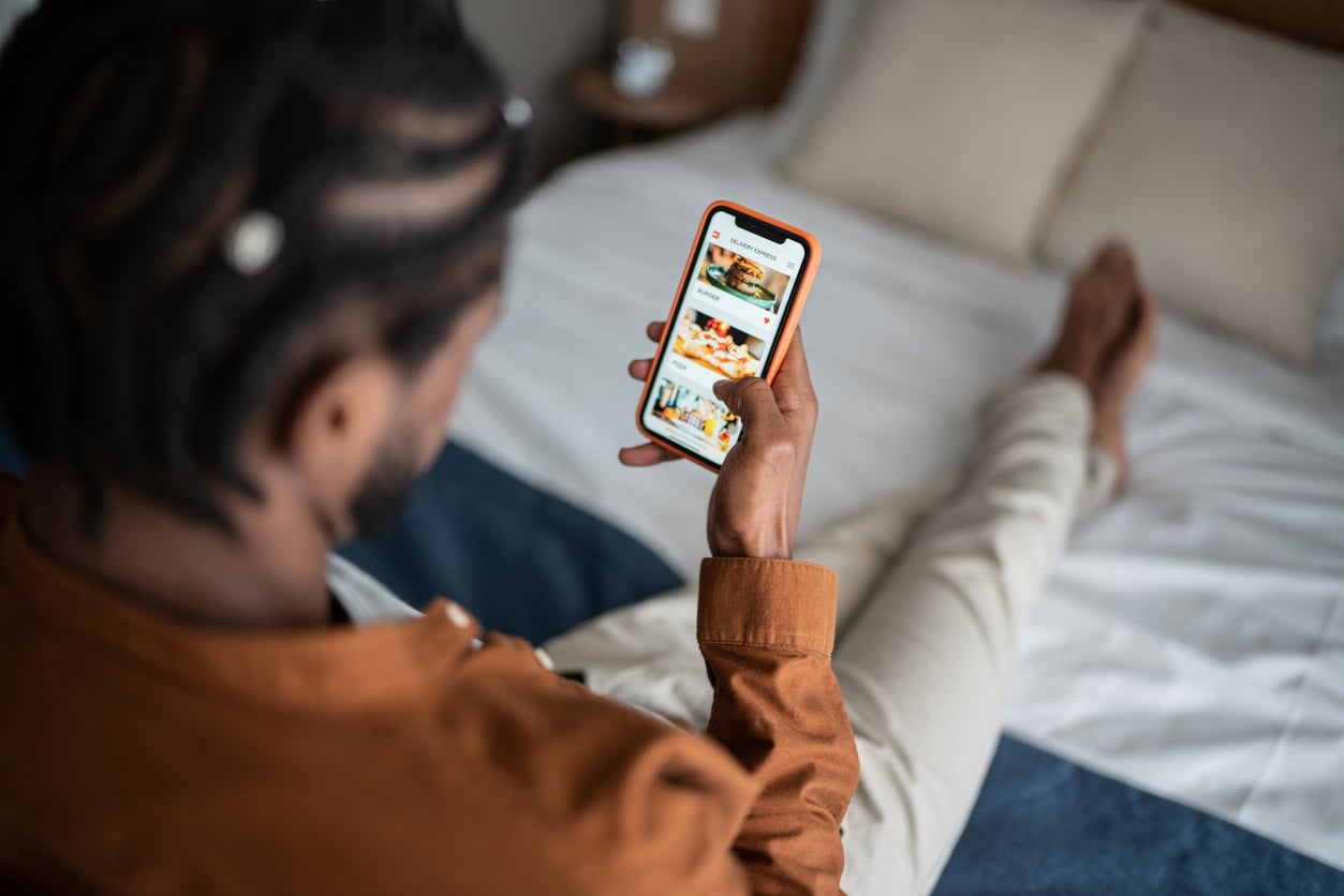 A person sitting on a bed looks at a smartphone displaying food delivery options on the screen, carefully considering money saving habits before placing an order.
