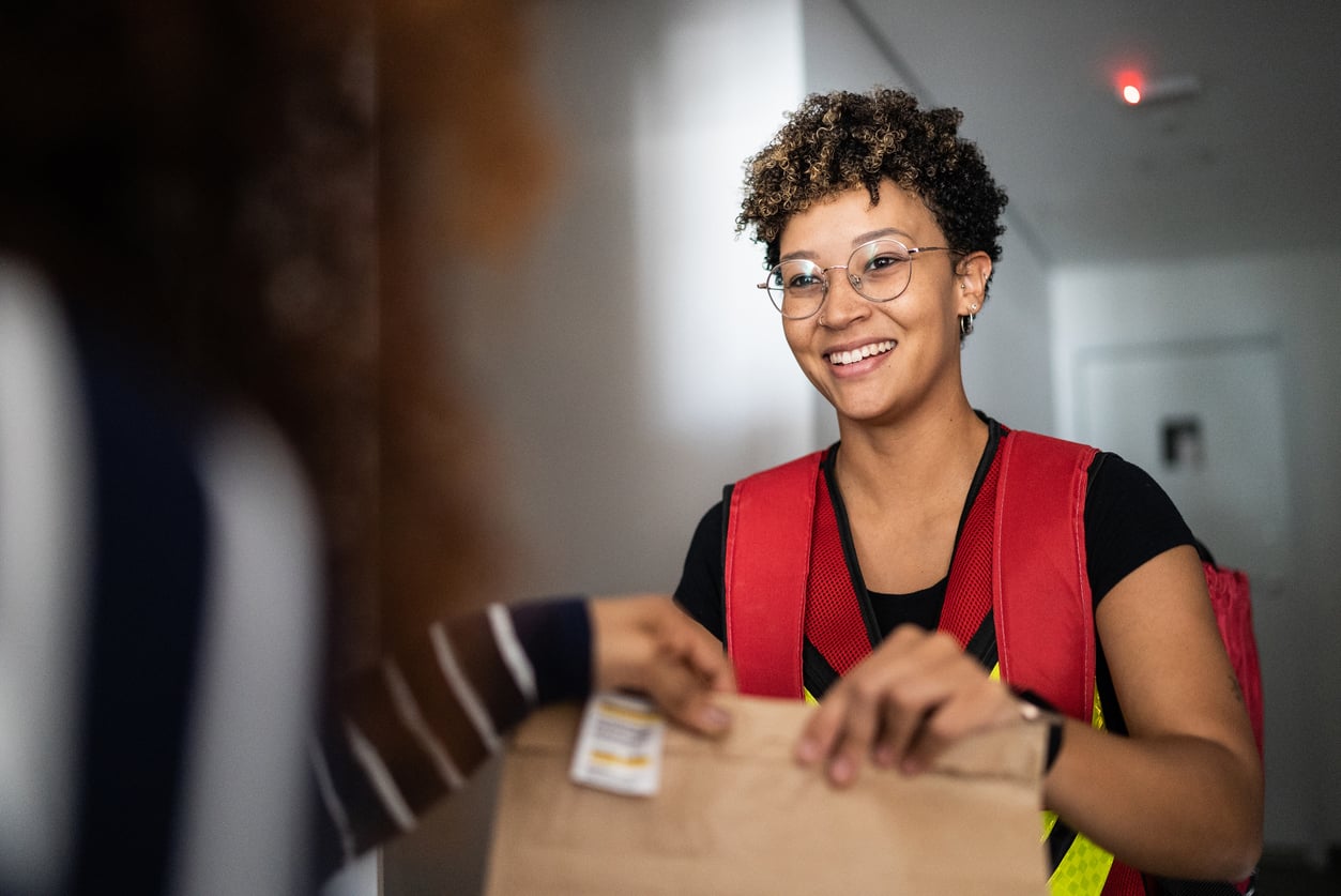 A smiling delivery worker in a safety vest hands a paper bag to a person indoors, with the recipient’s arm reaching out to take the package.